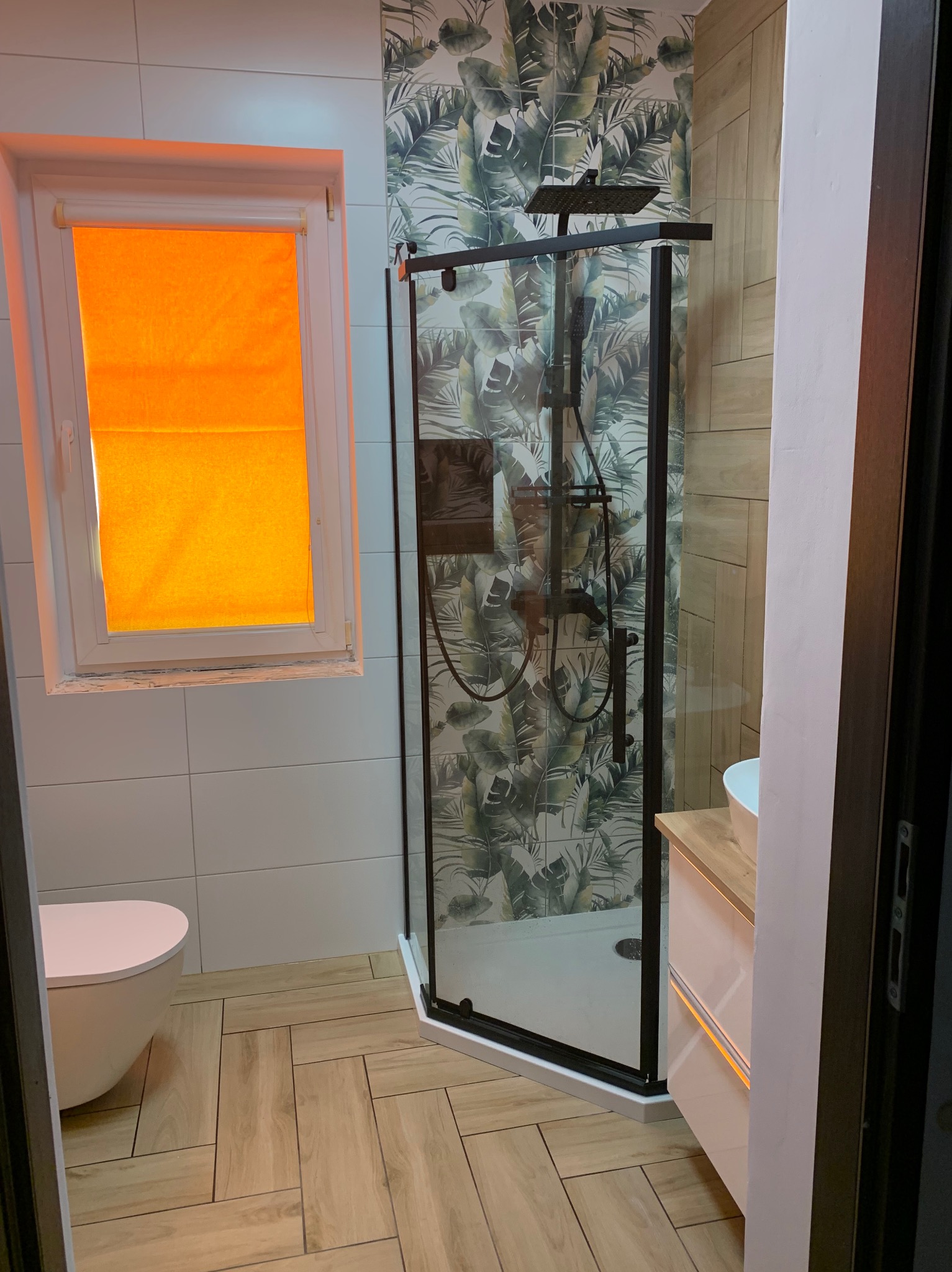 Nowoczesna łazienka z kabiną prysznicową typu walk-in z czarnymi profilami, umywalką z szafką, toaletą oraz oknem z pomarańczową roletą, wykończona płytkami imitującymi drewno i tapetą w motywy...