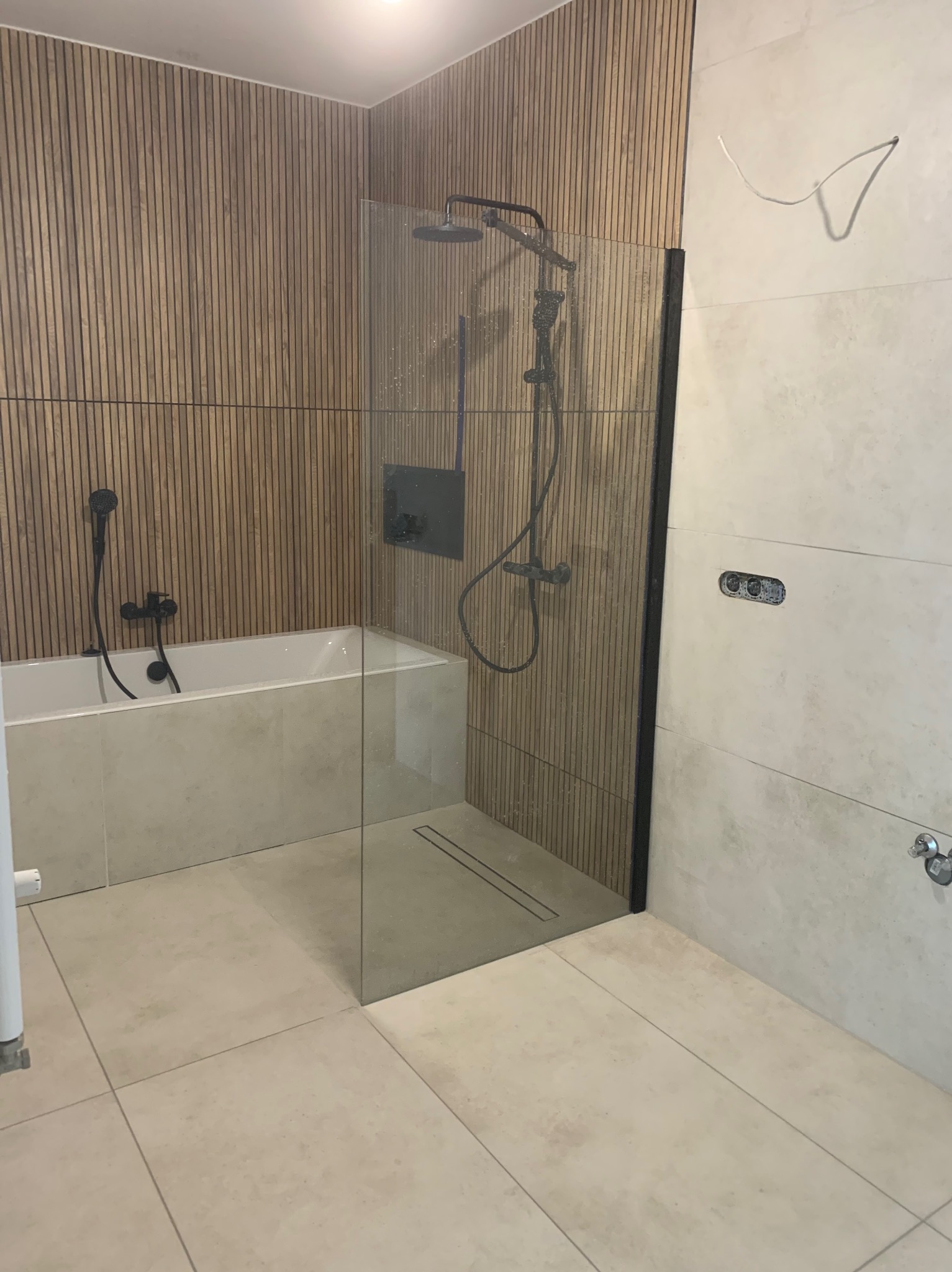 Nowoczesna łazienka z wanną obudowaną płytkami, kabiną prysznicową ze szklaną ścianką z czarnymi profilami, drewnianą okładziną ścienną i beżowymi płytkami podłogowymi, widoczne czarne baterie...