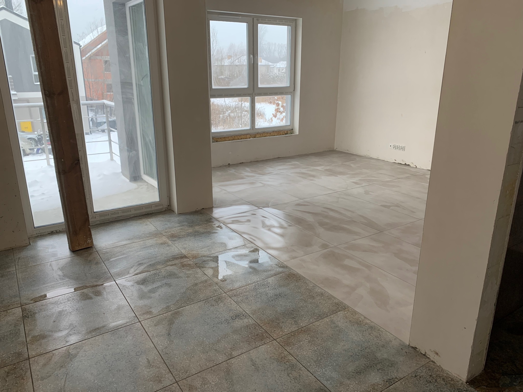 Wnętrze w stanie deweloperskim z nową, częściowo ułożoną podłogą z płytek imitujących kamień w odcieniach szarości i beżu, widok na okno z widokiem na zaśnieżone otoczenie budynku.