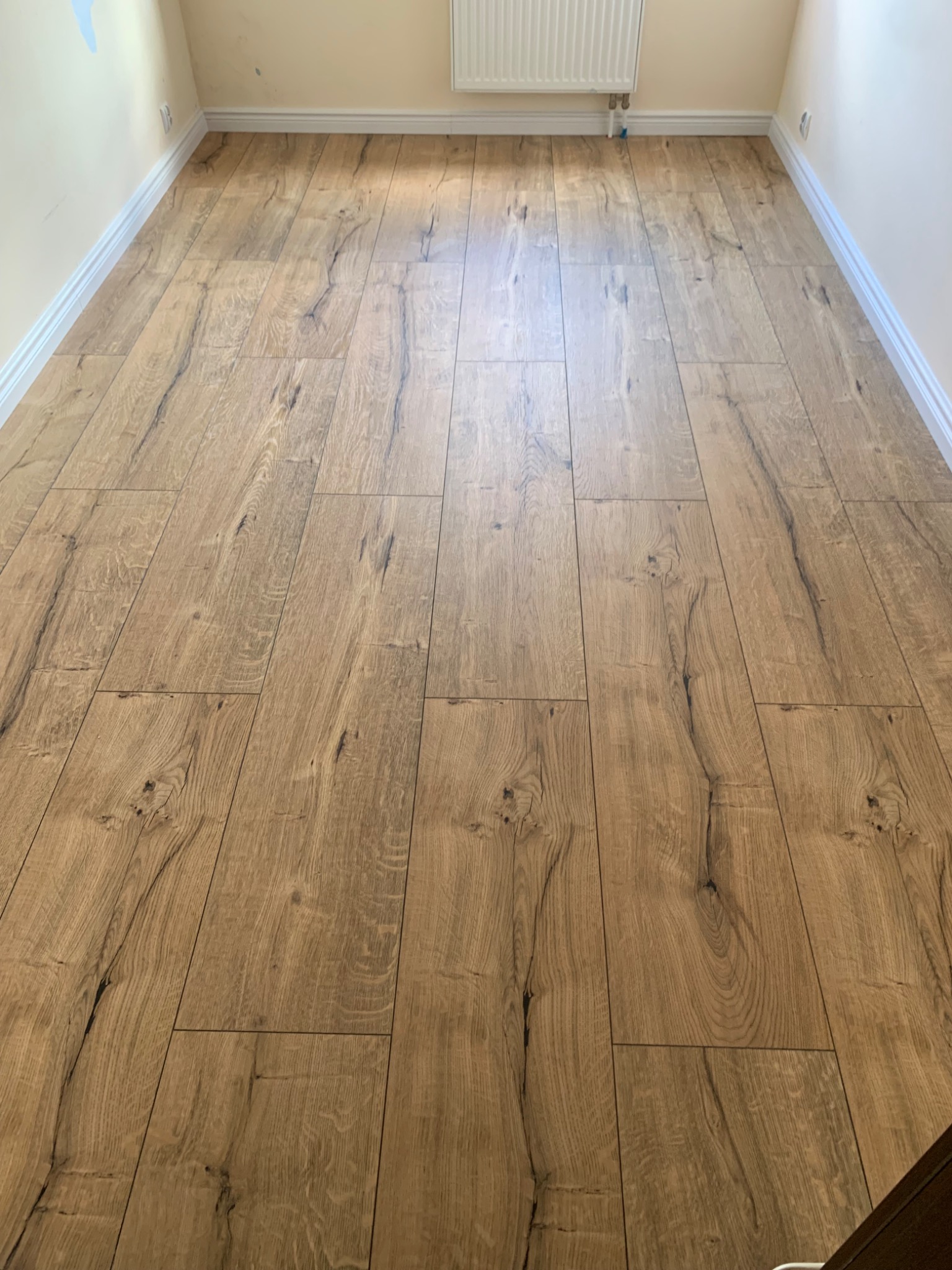Nowo ułożone panele podłogowe w odcieniach dębu, z wyraźnym rysunkiem słojów i subtelnymi czarnymi akcentami, wypełniają przestrzeń między białymi listwami przypodłogowymi i grzejnikiem.