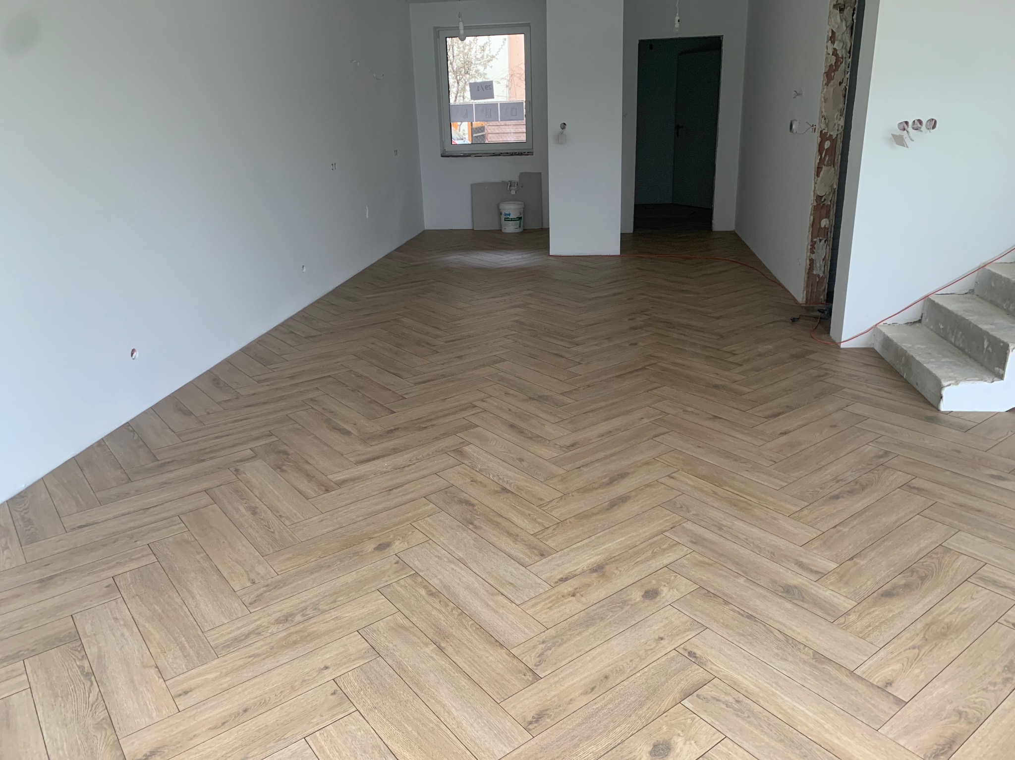 Jasne panele ułożone w jodełkę w przestronnym, surowym wnętrzu. Widoczne fragmenty nieotynkowanej ściany i surowe schody betonowe.