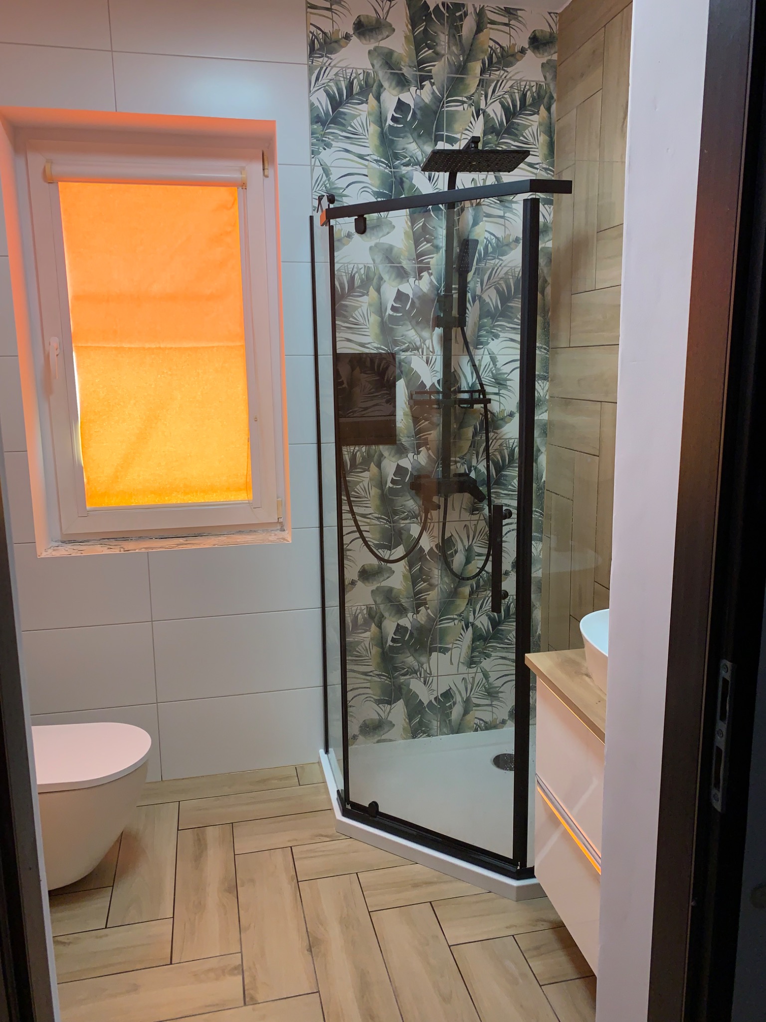Nowoczesna łazienka z kabiną prysznicową typu walk-in z czarną armaturą, drewnopodobne płytki na podłodze ułożone w jodełkę, ściana z motywem tropikalnych liści i pomarańczowa roleta w oknie.