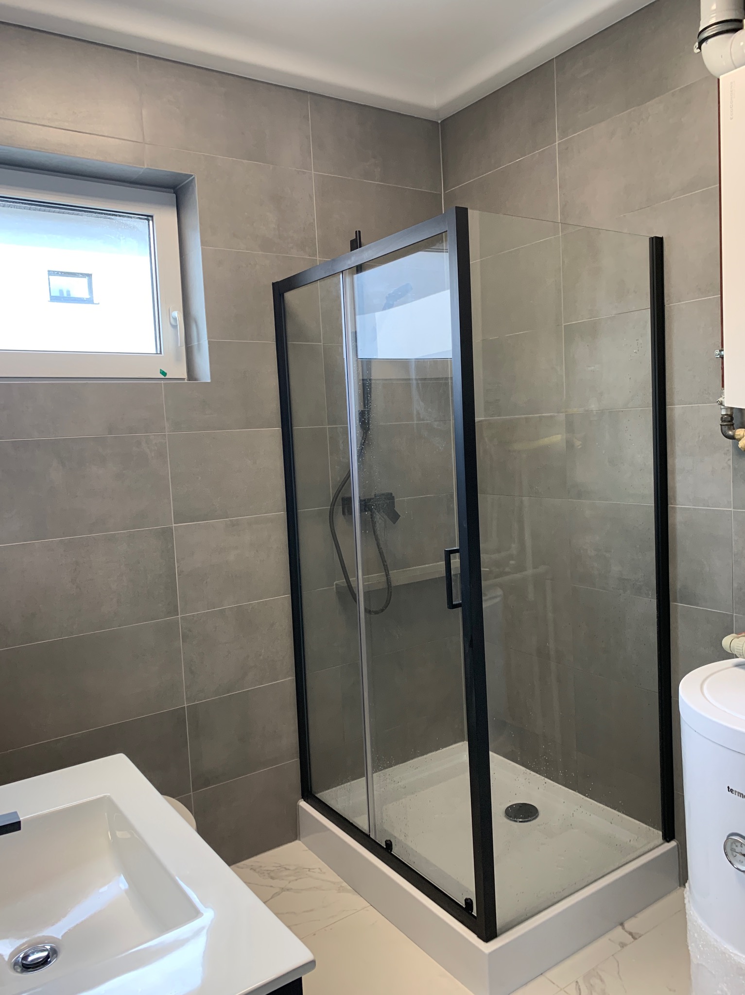 Nowoczesna kabina prysznicowa z czarnymi ramami i przesuwanymi drzwiami w łazience wyłożonej szarymi płytkami, widoczna biała umywalka i okno.