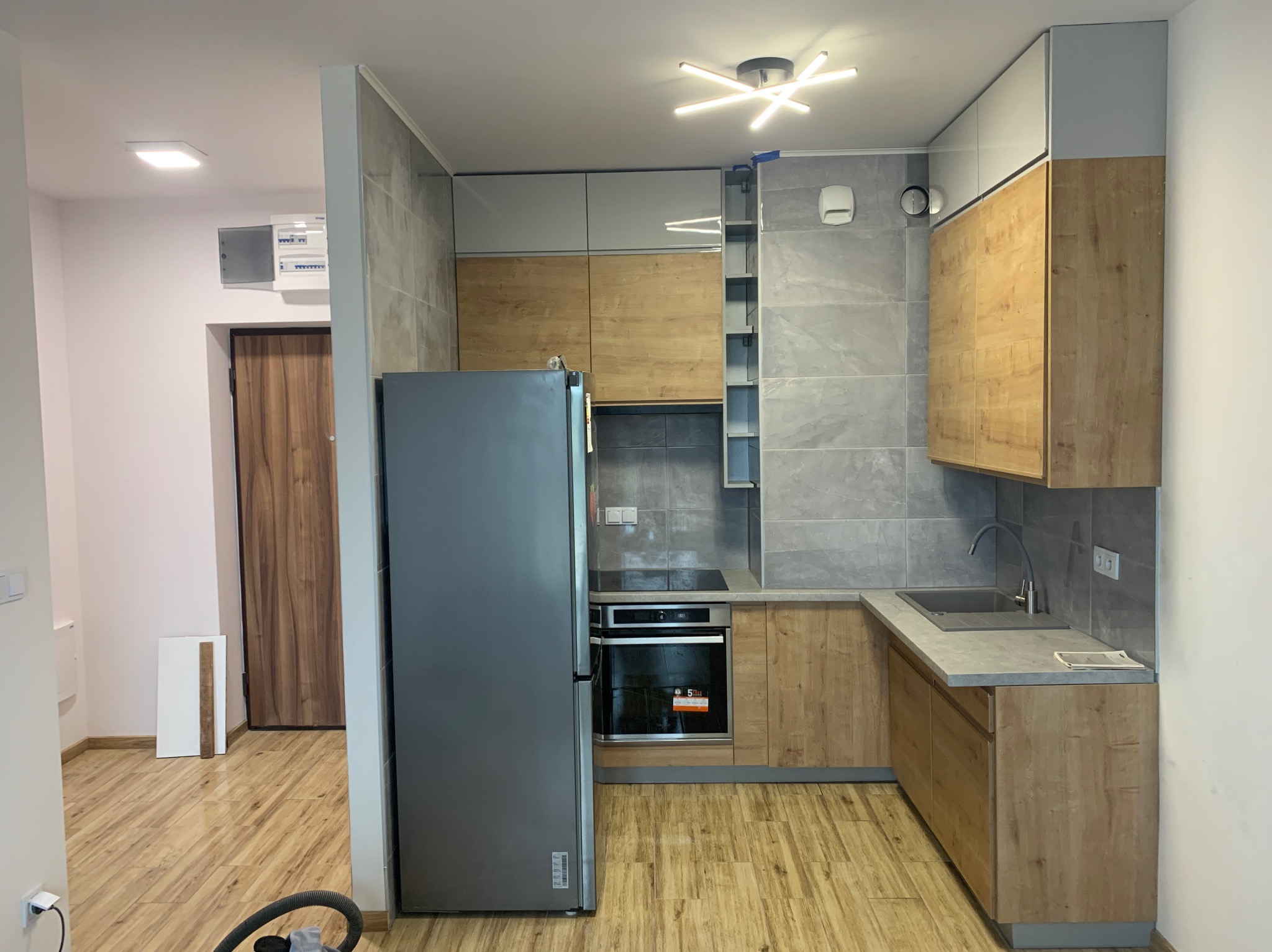 Nowoczesna kuchnia z szarymi płytkami i drewnopodobnymi frontami szafek, srebrna lodówka, oświetlenie sufitowe w kształcie gwiazdy, widok na korytarz z drewnianą podłogą i drzwiami.