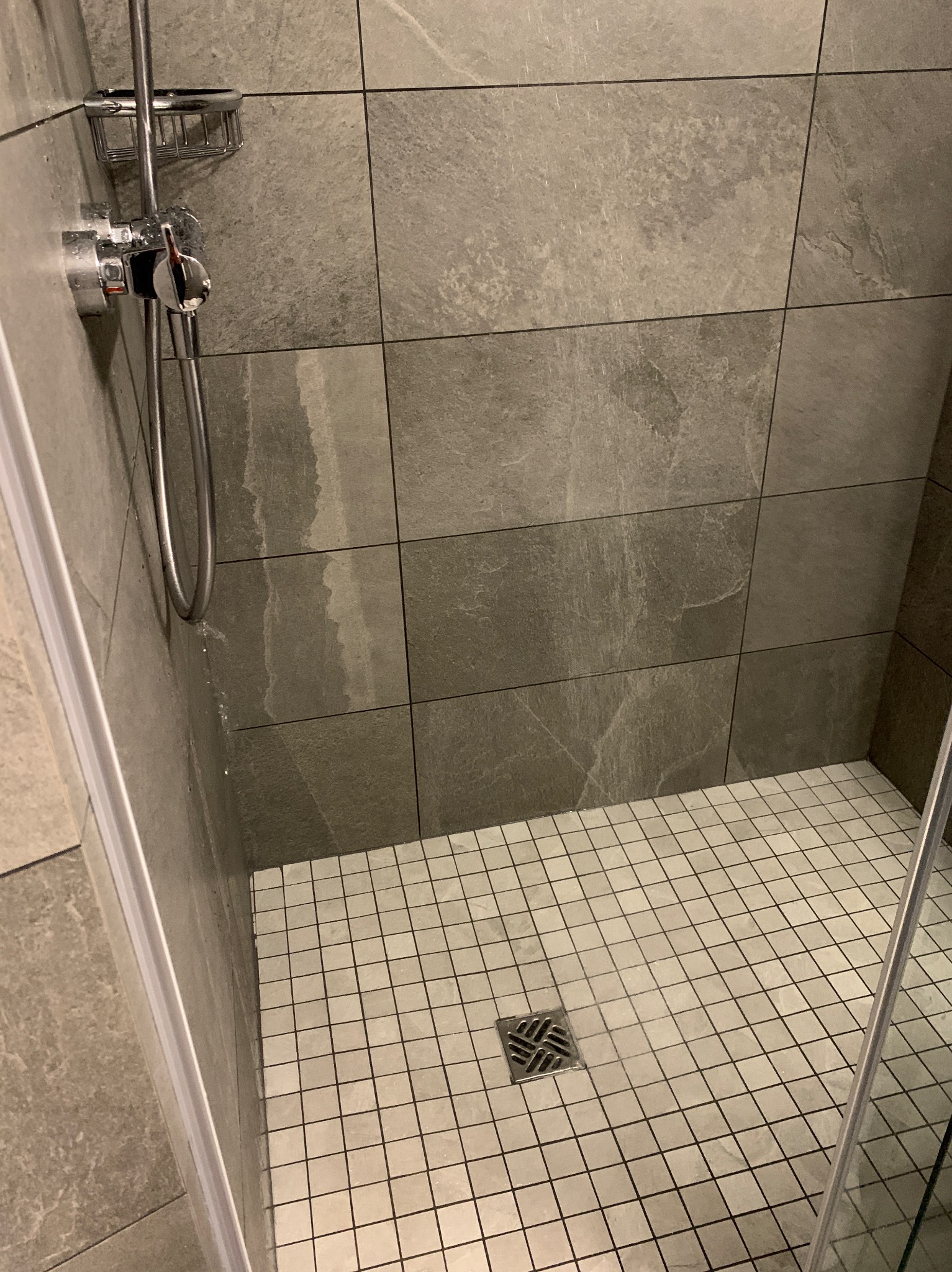 Nowoczesny prysznic z szarymi płytkami imitującymi kamień na ścianach i mozaiką na podłodze, widoczny chromowany zestaw natryskowy z mydelniczką i odpływ liniowy.