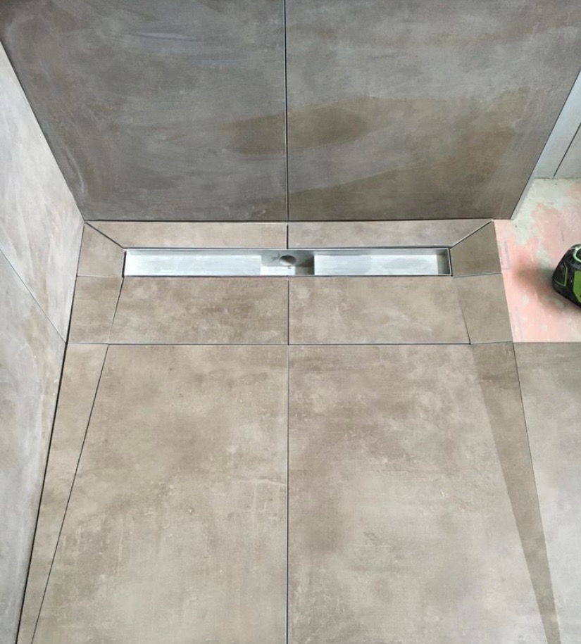 Ułożone płytki podłogowe w łazience z odpływem liniowym w trakcie prac wykończeniowych, widoczny laser krzyżowy na różowym podkładzie podłogowym.