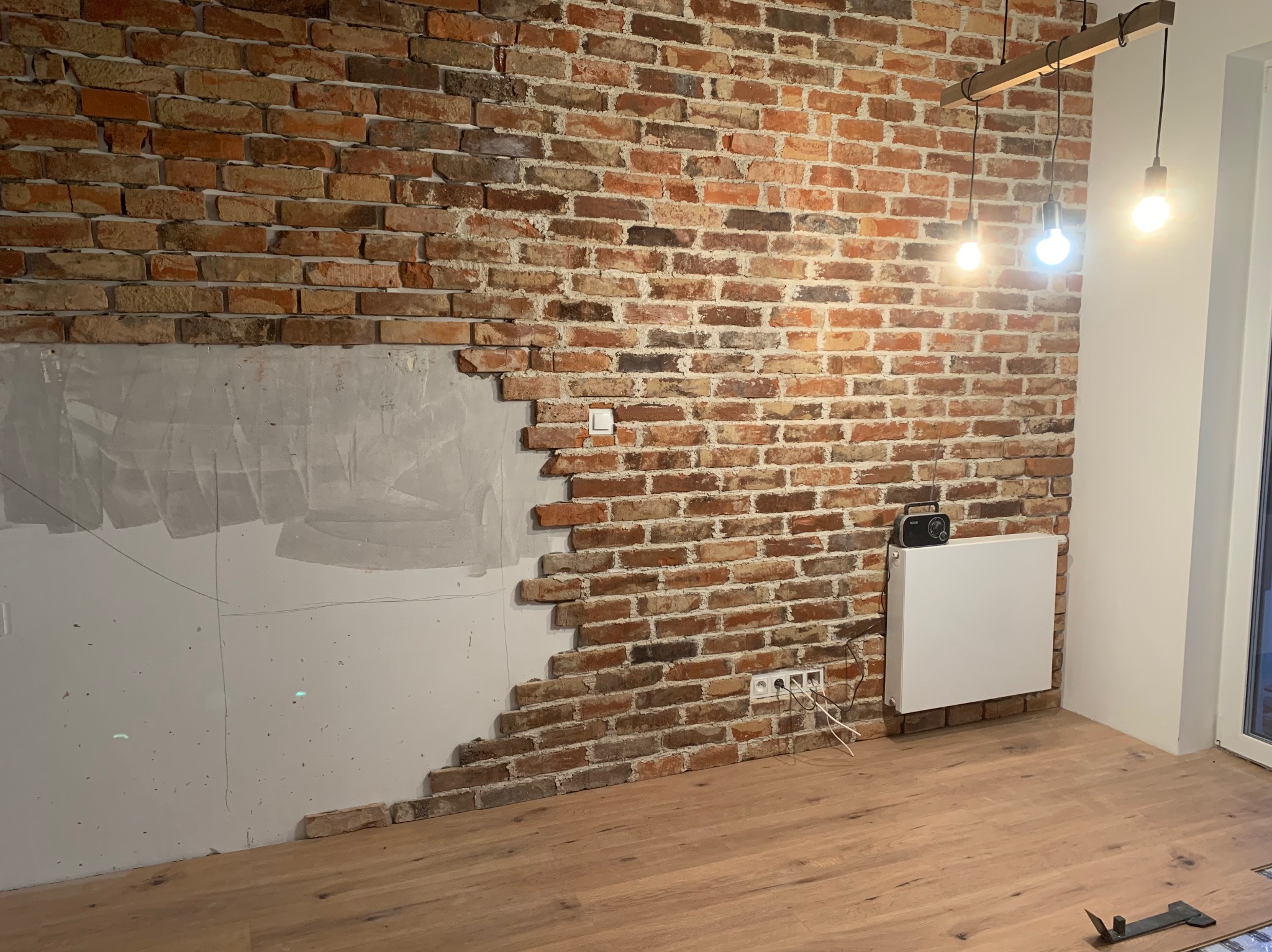 Częściowo otynkowana ceglana ściana z widocznymi gniazdkami elektrycznymi, grzejnikiem i radiem, na podłodze ułożone panele, surowa lampa wisząca z trzema żarówkami