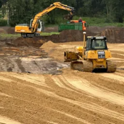 Żółta spycharka wyrównuje teren piaszczysty, w tle koparka Liebherr załadowuje ziemię na ciężarówkę, widoczne ślady opon na piasku.