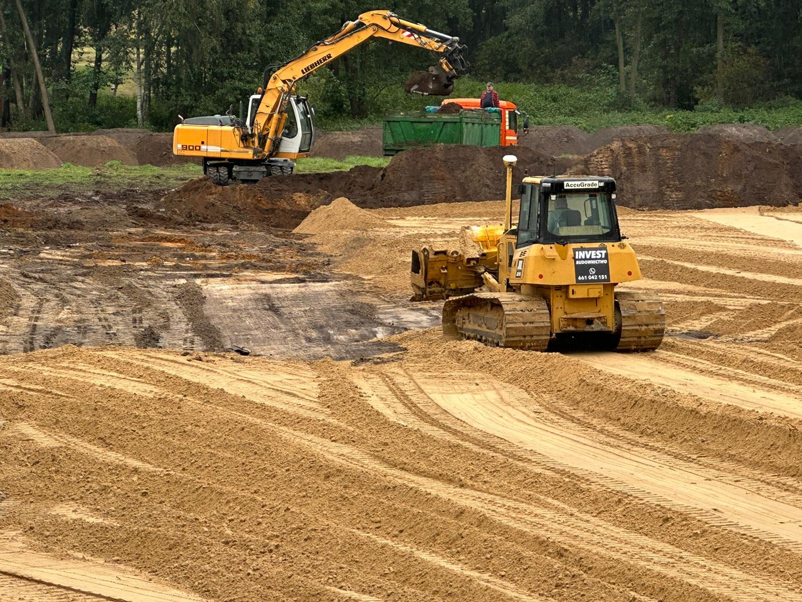 Żółta spycharka wyrównuje teren piaszczysty, w tle koparka Liebherr załadowuje ziemię na ciężarówkę, widoczne ślady opon na piasku.