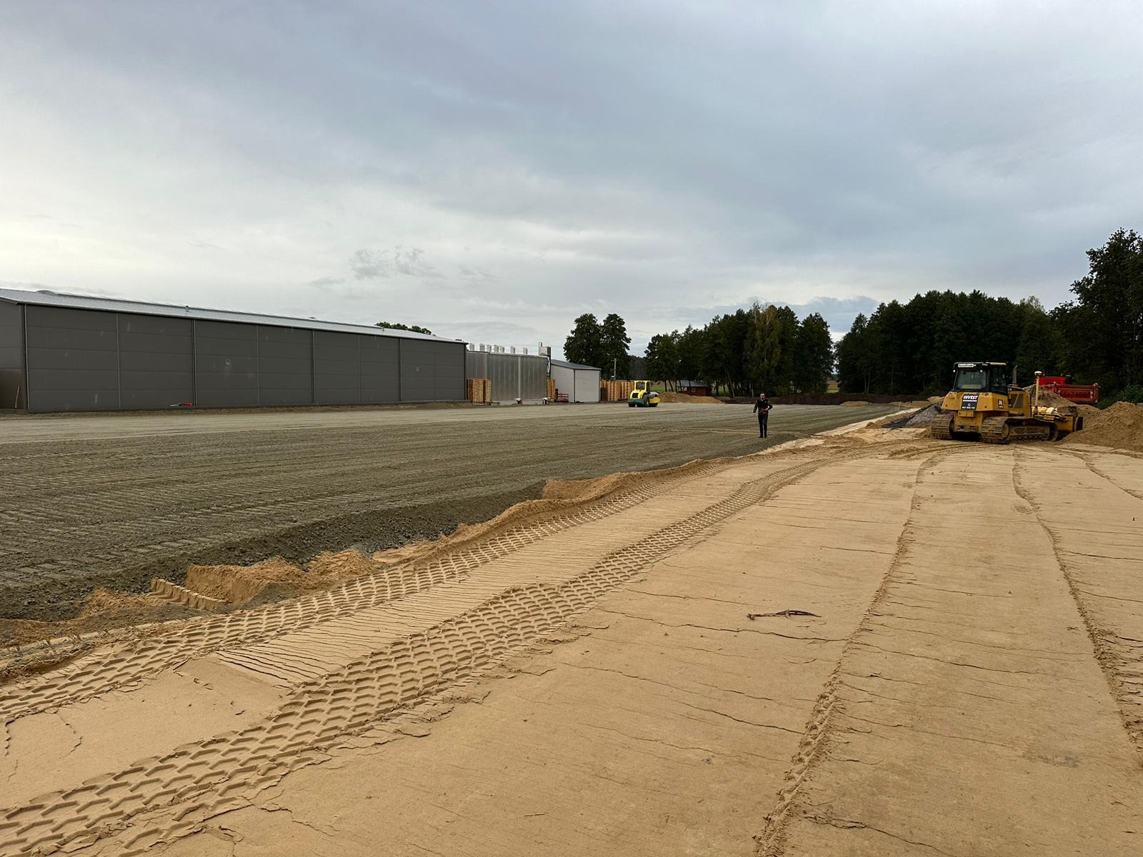 Przygotowanie terenu pod budowę hali: wyrównywanie gruntu i ubijanie walcem, widoczny spychacz i pracownik nadzorujący.