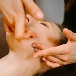Masaż twarzy wykonywany na leżącej kobiecie, skupienie na dłoniach masażysty i skórze twarzy klientki.