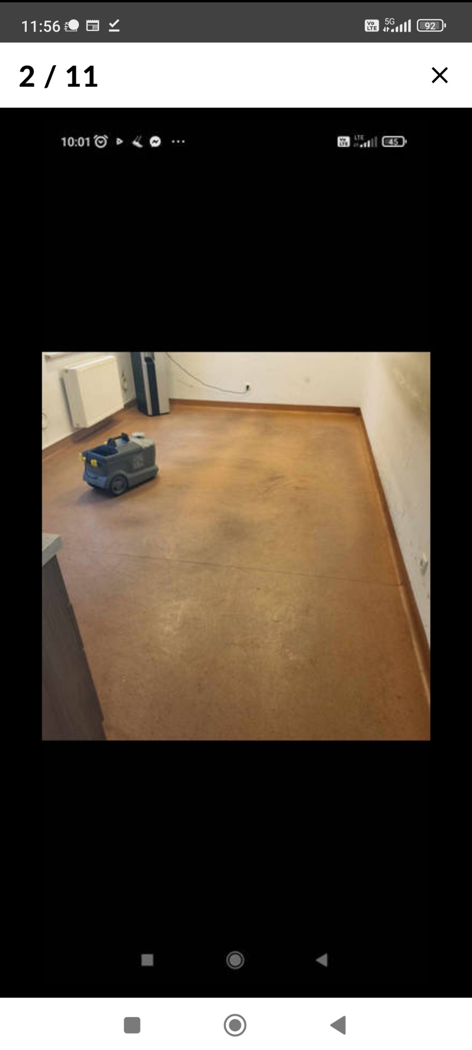 Szorowarka do podłóg na tle świeżo umytej, brązowej podłogi w pomieszczeniu z grzejnikiem i listwami przypodłogowymi.