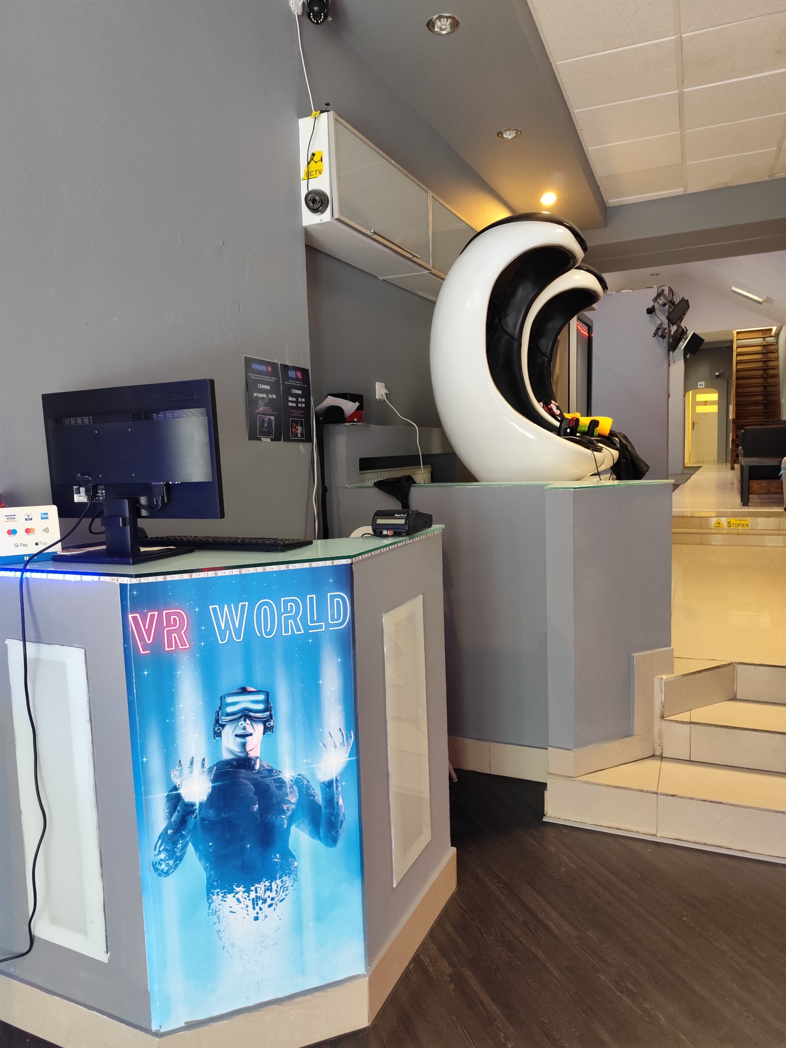 Wnętrze salonu VR World z futurystycznym fotelem do wirtualnej rzeczywistości, ladą z podświetlanym logo oraz monitorem z cennikiem.