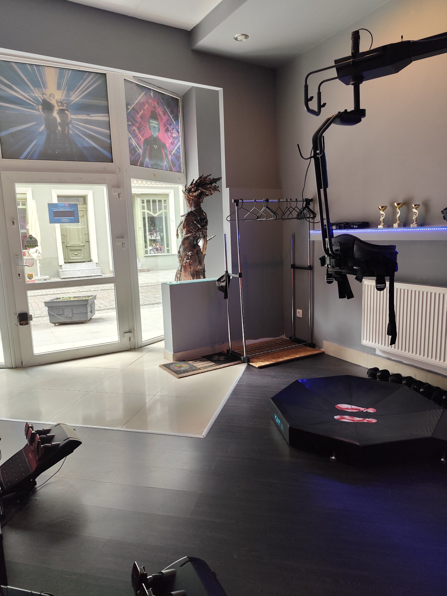 Wnętrze studia z platformą do wirtualnej rzeczywistości, widoczne plakaty na ścianach, rzeźba i trofea na półce.