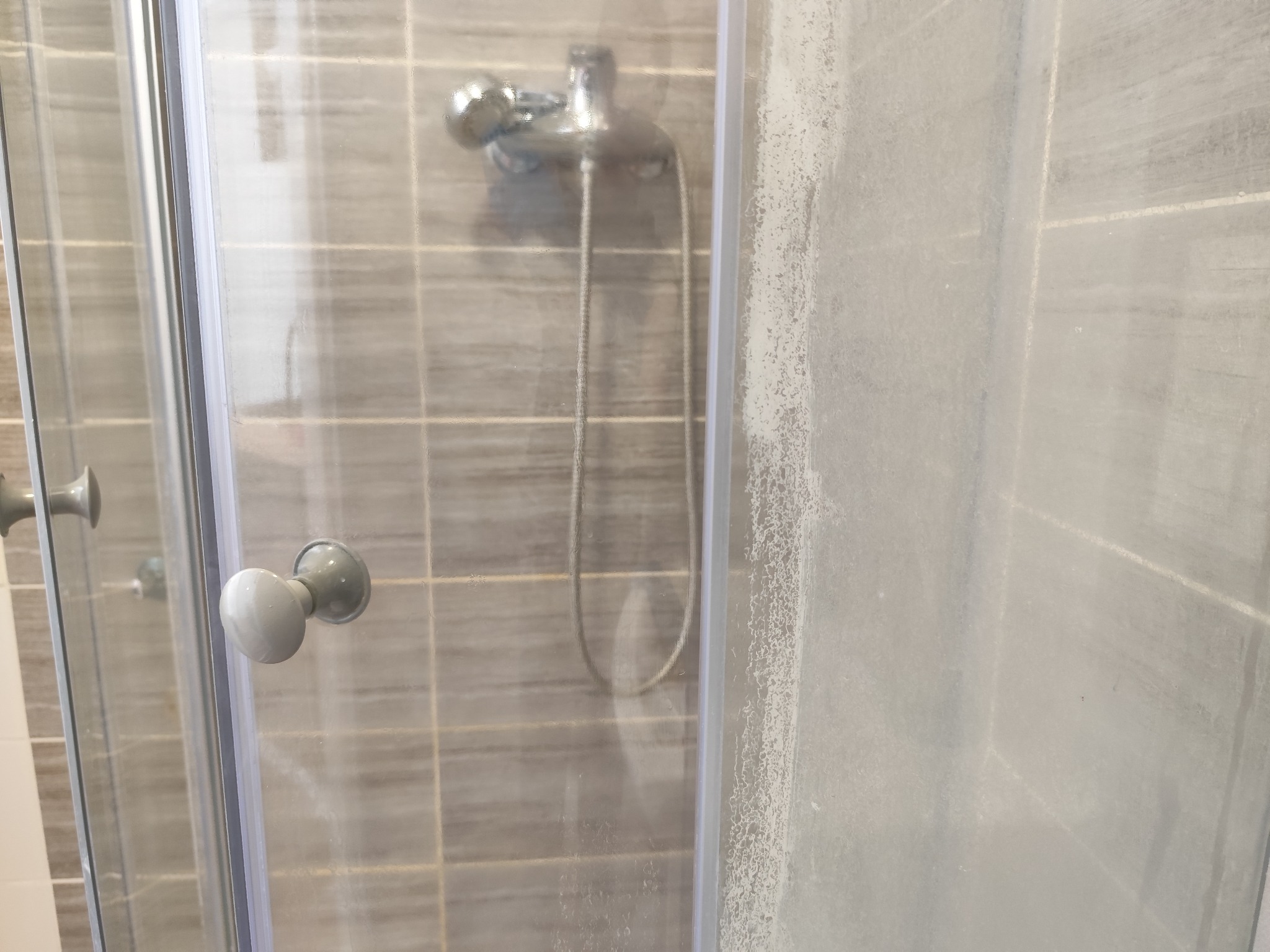 Kabina prysznicowa z widocznymi śladami kamienia i mydła na szklanych drzwiach, chromowana bateria i słuchawka prysznicowa na tle szarych płytek.