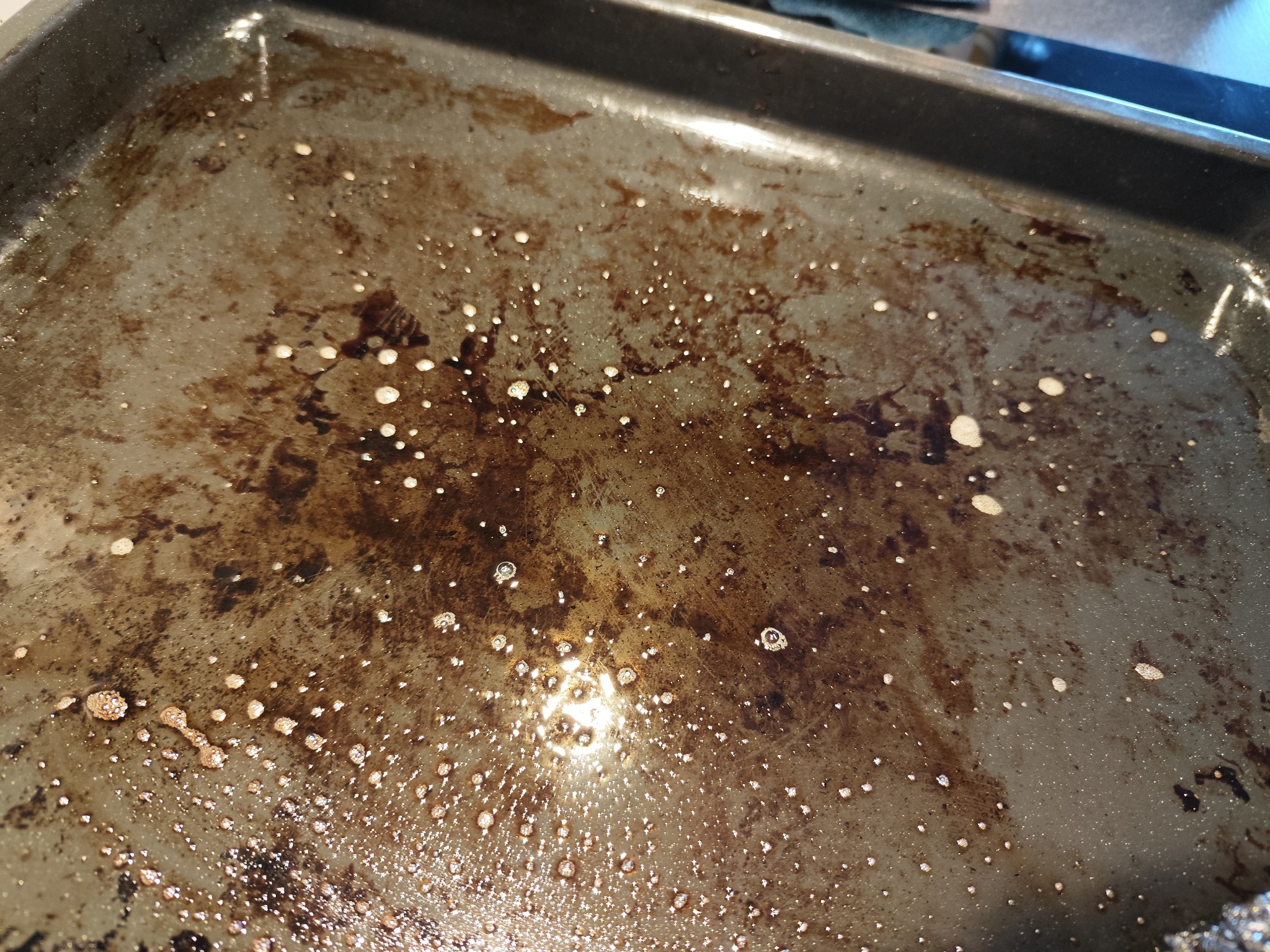 Brudna blacha do pieczenia z przypalonym tłuszczem i resztkami jedzenia, widoczne krople wody po próbie czyszczenia.