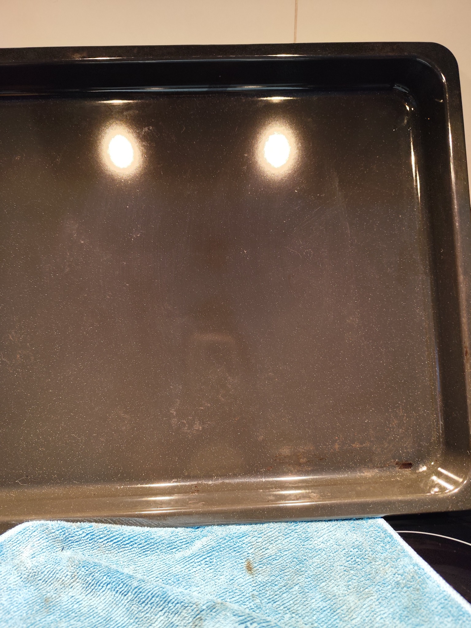 Brudna, prostokątna blacha do pieczenia, częściowo umyta, leżąca na niebieskiej ściereczce z mikrofibry.