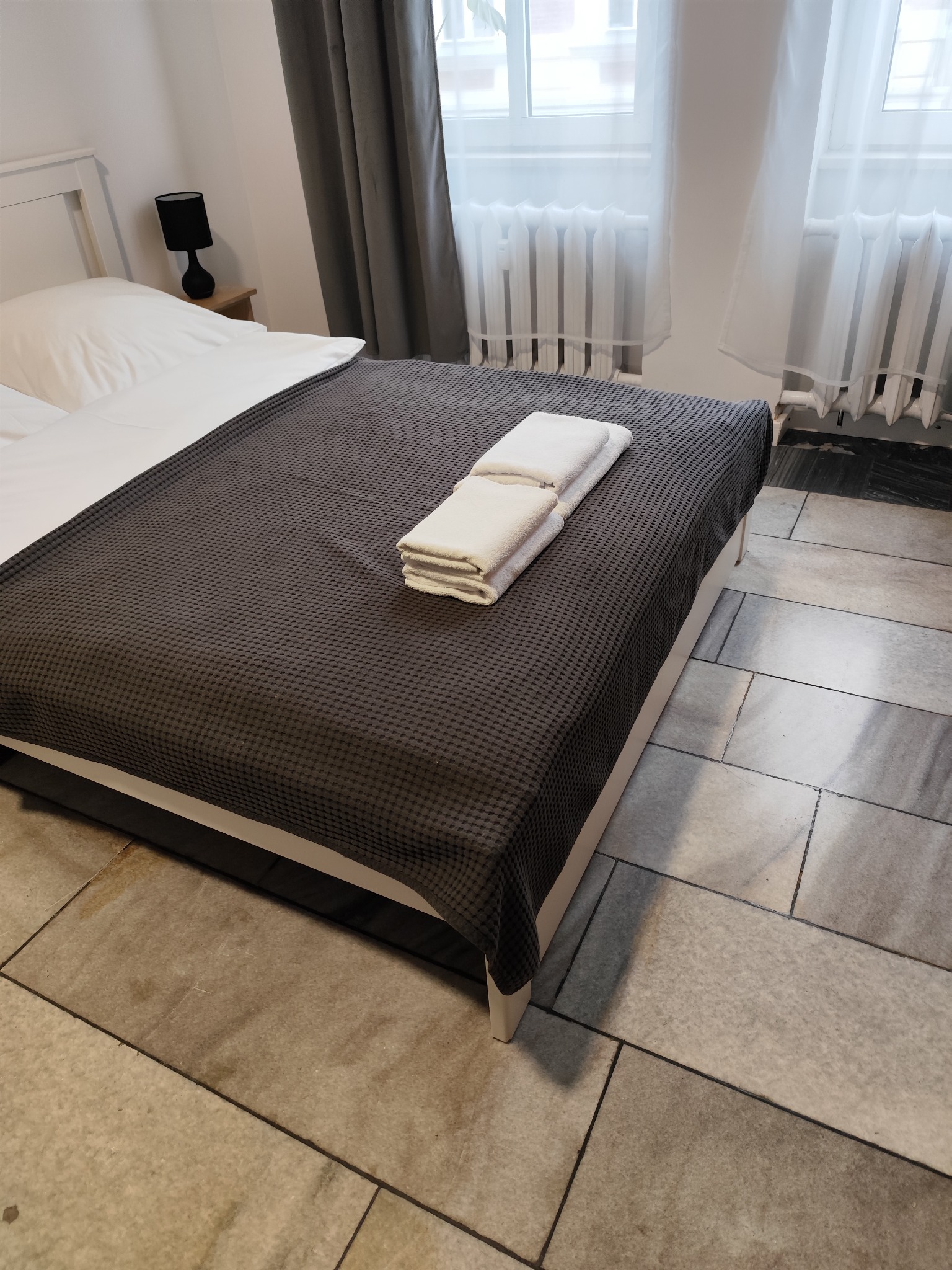 Przygotowane łóżko w pokoju hotelowym z szarą narzutą i stertą białych ręczników, widoczne płytki podłogowe i fragment grzejnika pod oknem.