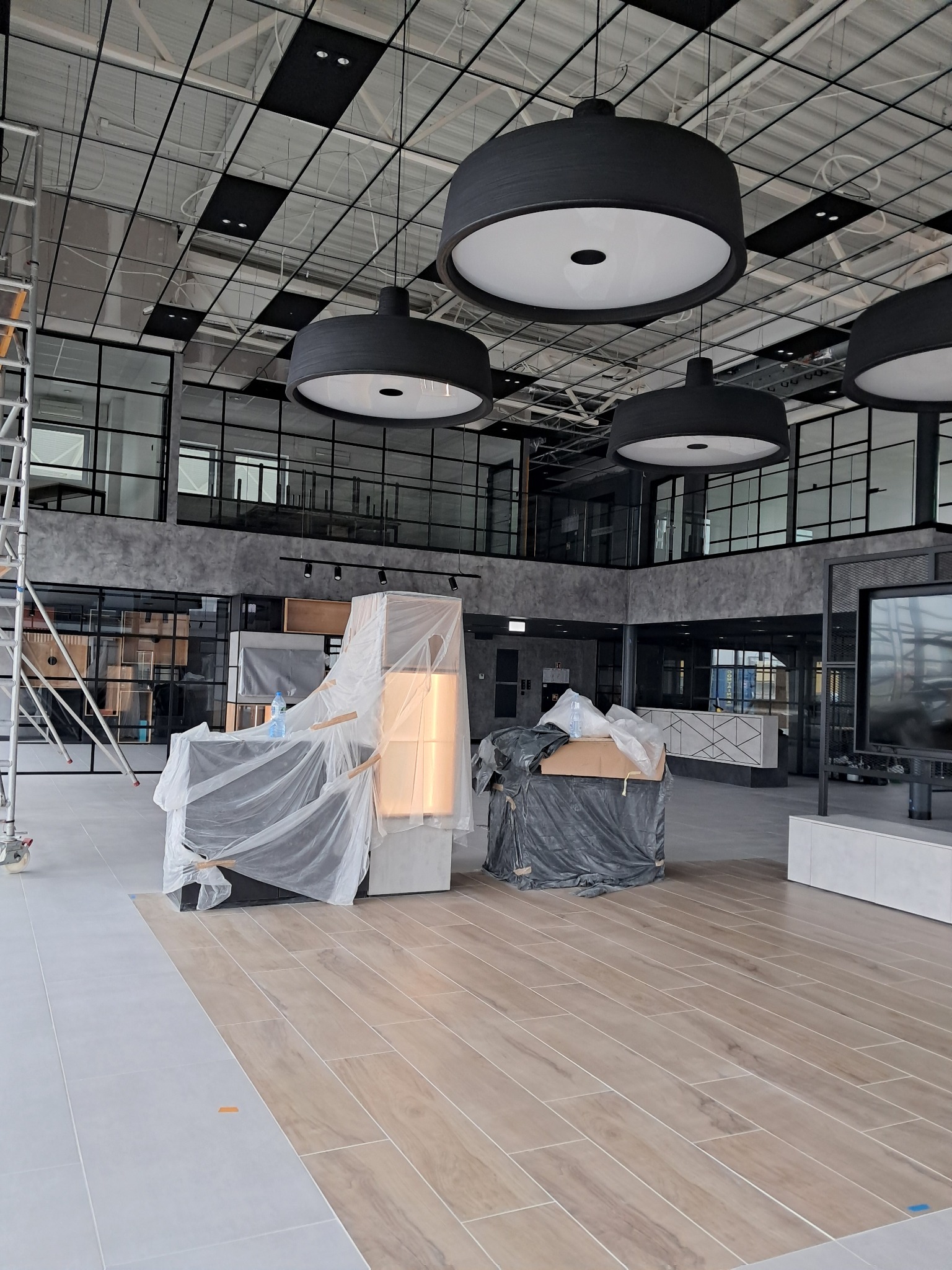 Nowoczesne biuro w trakcie wykańczania: meble zabezpieczone folią, surowy sufit z lampami, częściowo ułożona podłoga.