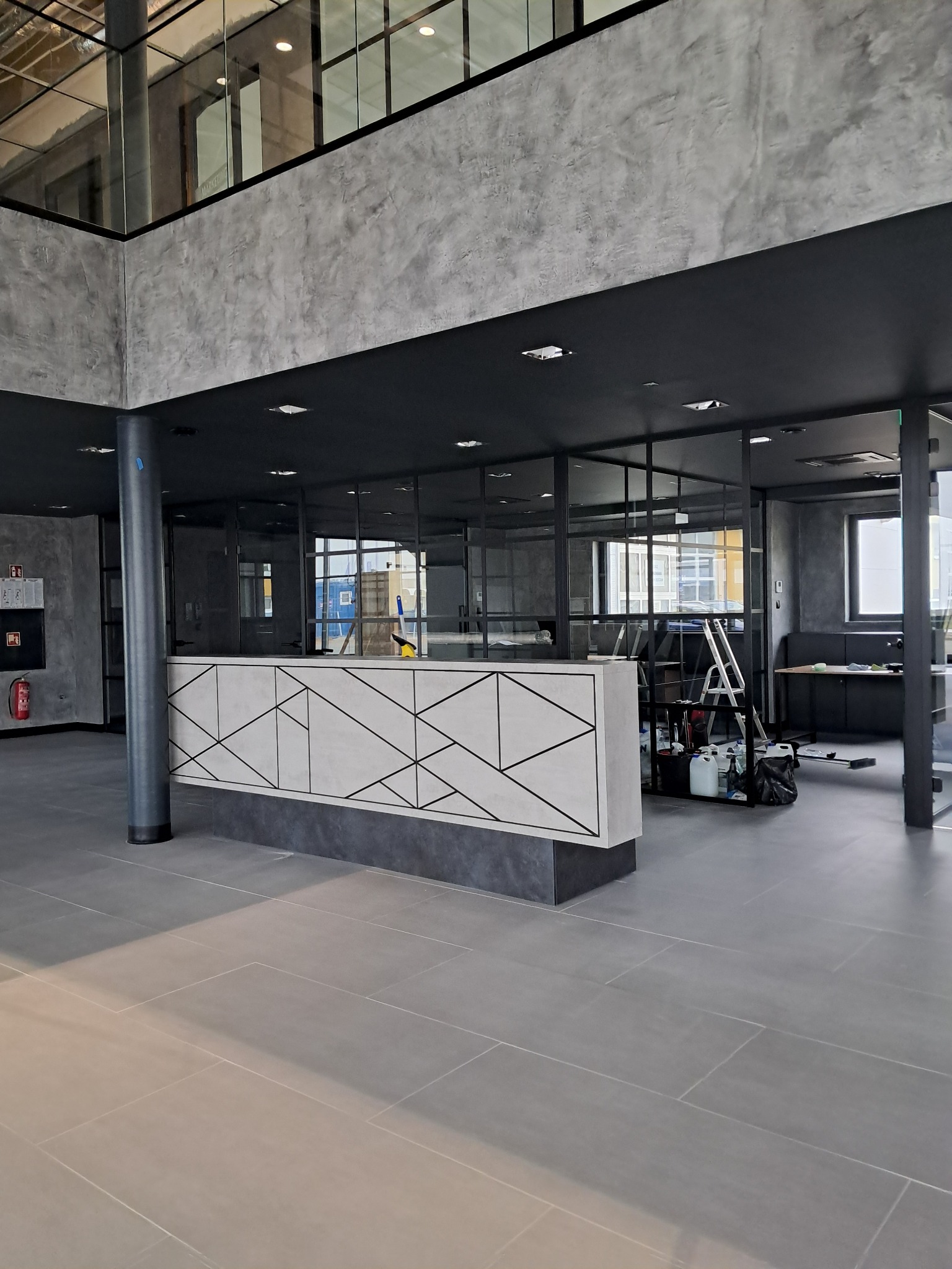 Wnętrze biurowe z recepcją o geometrycznym wzorze, widoczne butelki i drabina sugerujące prace porządkowe w toku.