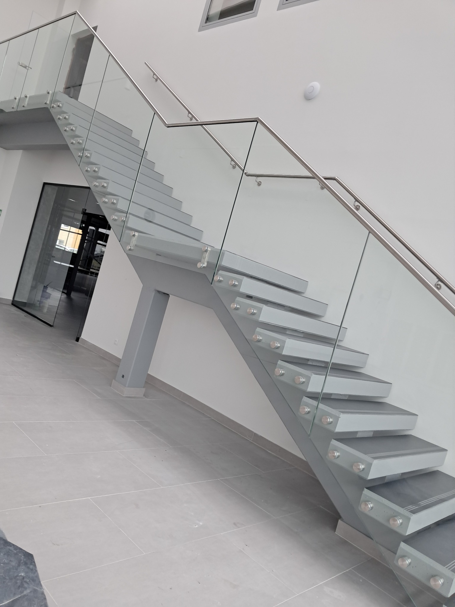 Nowoczesne, szare schody z szklanymi balustradami i metalowymi poręczami w jasnym wnętrzu biurowym.