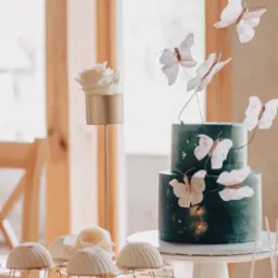 Dwupiętrowy tort w odcieniach zieleni, zdobiony motylami i złotymi płatkami, ustawiony na białym stojaku. Obok desery w kształcie kopuł na złotej kratce oraz biała r&oacute;ża w złotym, wysokim wazonie.