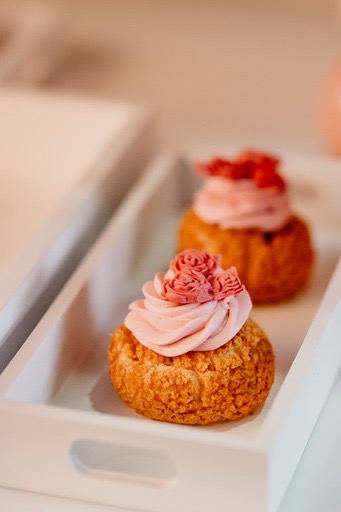 Dwa ptysie na białej, drewnianej tacy. Pierwszy plan: ptys z kremem w odcieniach różu uformowanym w róże. Drugi plan: ptys z kremem i drobnymi owocami.