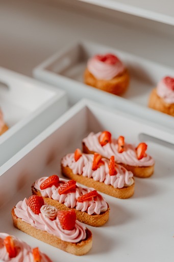 Rząd podłużnych tartaletek z różowym kremem i owocami (maliny, truskawki) na białych, drewnianych tacach; w tle pojedyncze babeczki z różowym kremem.