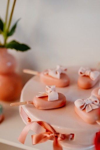 Różowe cake pops w kształcie serc z kokardkami na różowym paterze, dekorowane wstążką, z wazonem z kwiatami w tle.