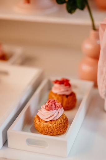 Dwa ciastka typu ptysie na białych prostokątnych tackach. Pierwsze ciastko z kremem w kolorze różowym uformowanym w różyczki, drugie z kremem i małymi czerwonymi owocami. W tle pastelowe dekoracje.