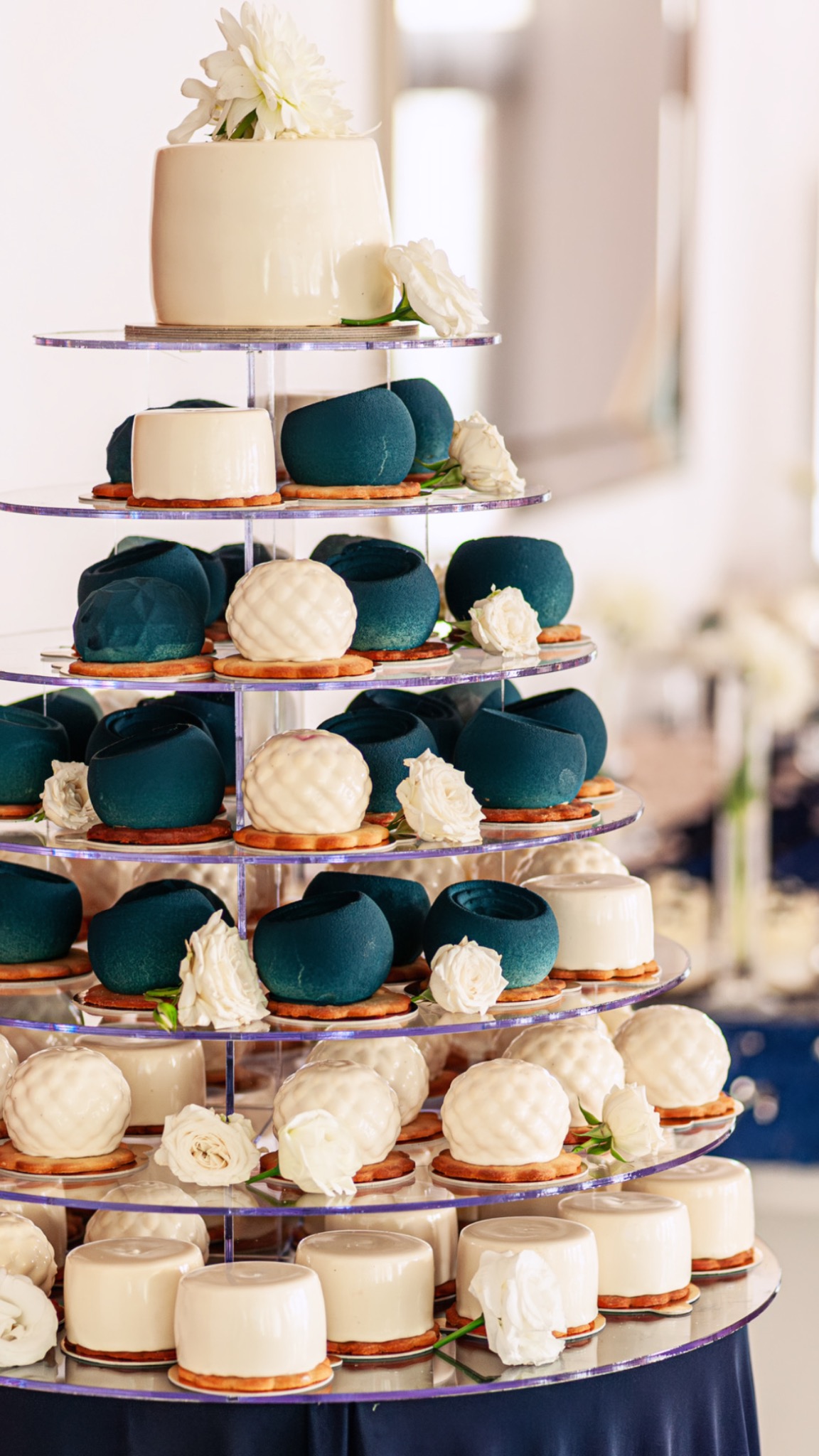 Wielopoziomowa ekspozycja deserów: białe mini-tortowe kopułki, kule z teksturą oraz turkusowe musowe babeczki, udekorowane białymi różami, na przezroczystych półkach.