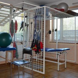 SPECJALISTYCZNY ZAKŁAD REHABILITACJI PRĄDNIK SPÓŁKA Z OGRANICZONĄ ODPOWIEDZIALNOŚCIĄ - Sala rehabilitacyjna z drabinką rehabilitacyjną, stołem do ćwiczeń i piłkami gimnastycznymi, oświetlona przez okno z żaluzjami.