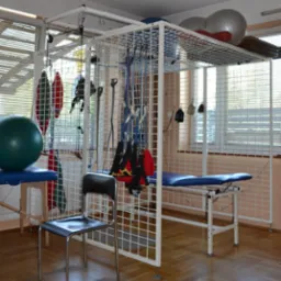 Sala rehabilitacyjna z drabinką rehabilitacyjną, stołem do ćwiczeń i piłkami gimnastycznymi, oświetlona przez okno z żaluzjami.