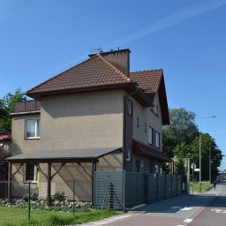 SPECJALISTYCZNY ZAKŁAD REHABILITACJI PRĄDNIK SPÓŁKA Z OGRANICZONĄ ODPOWIEDZIALNOŚCIĄ - Dwupiętrowy dom z beżową elewacją i brązowym dachem, widoczny z perspektywy ulicy, z przylegającym zadaszeniem i ogrodzeniem z poziomych desek.