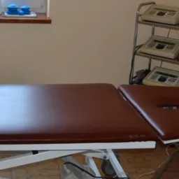 Brązowy stół do terapii z otworem na twarz, obok metalowy wózek z trzema urządzeniami do elektroterapii, w tle okno z niebieskimi bańkami do masażu.