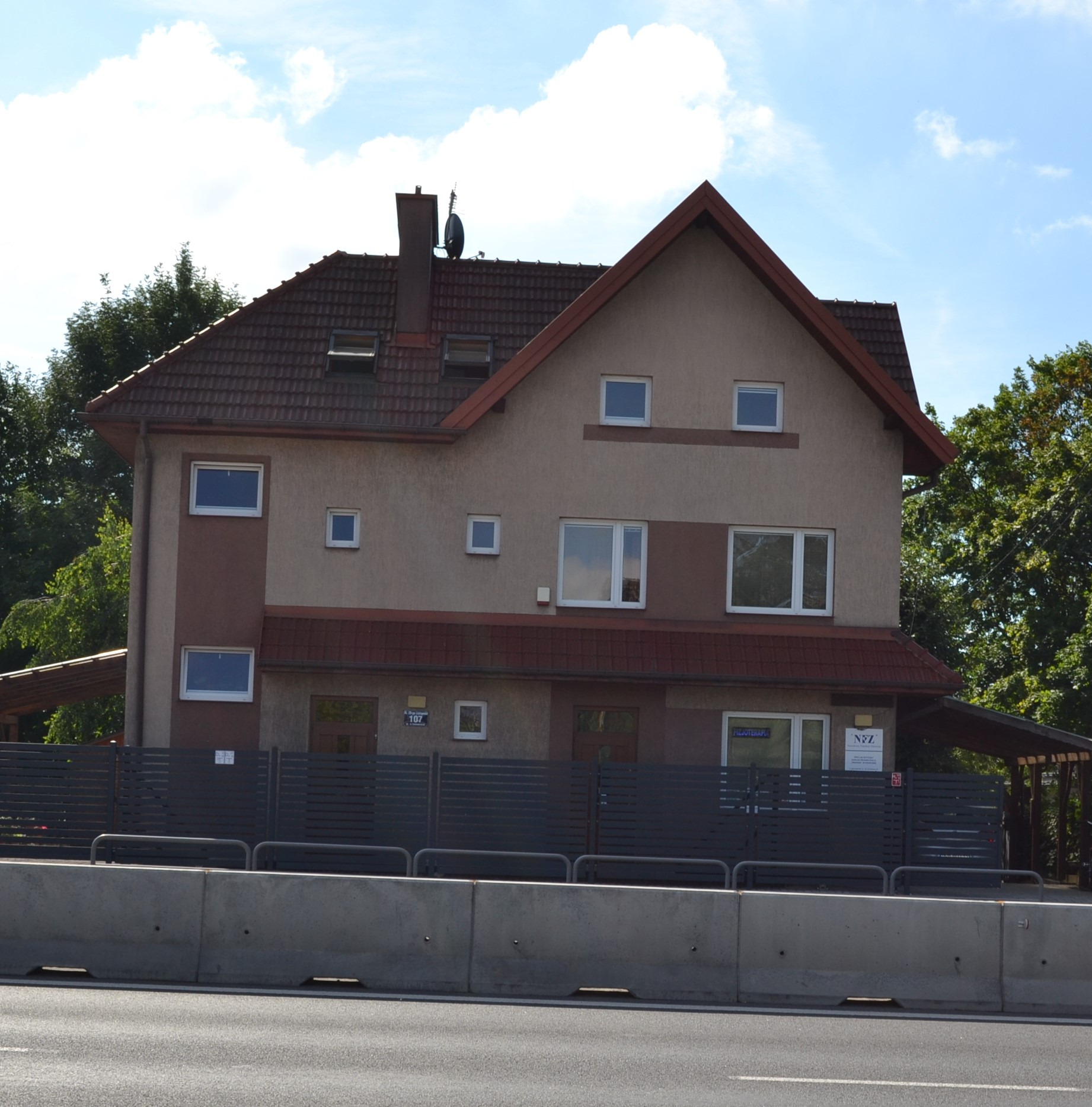 Dwupiętrowy budynek z brązową dachówką i beżową elewacją, widoczny z ulicy, z tabliczką informacyjną 'Fizjoterapia' w oknie na parterze.