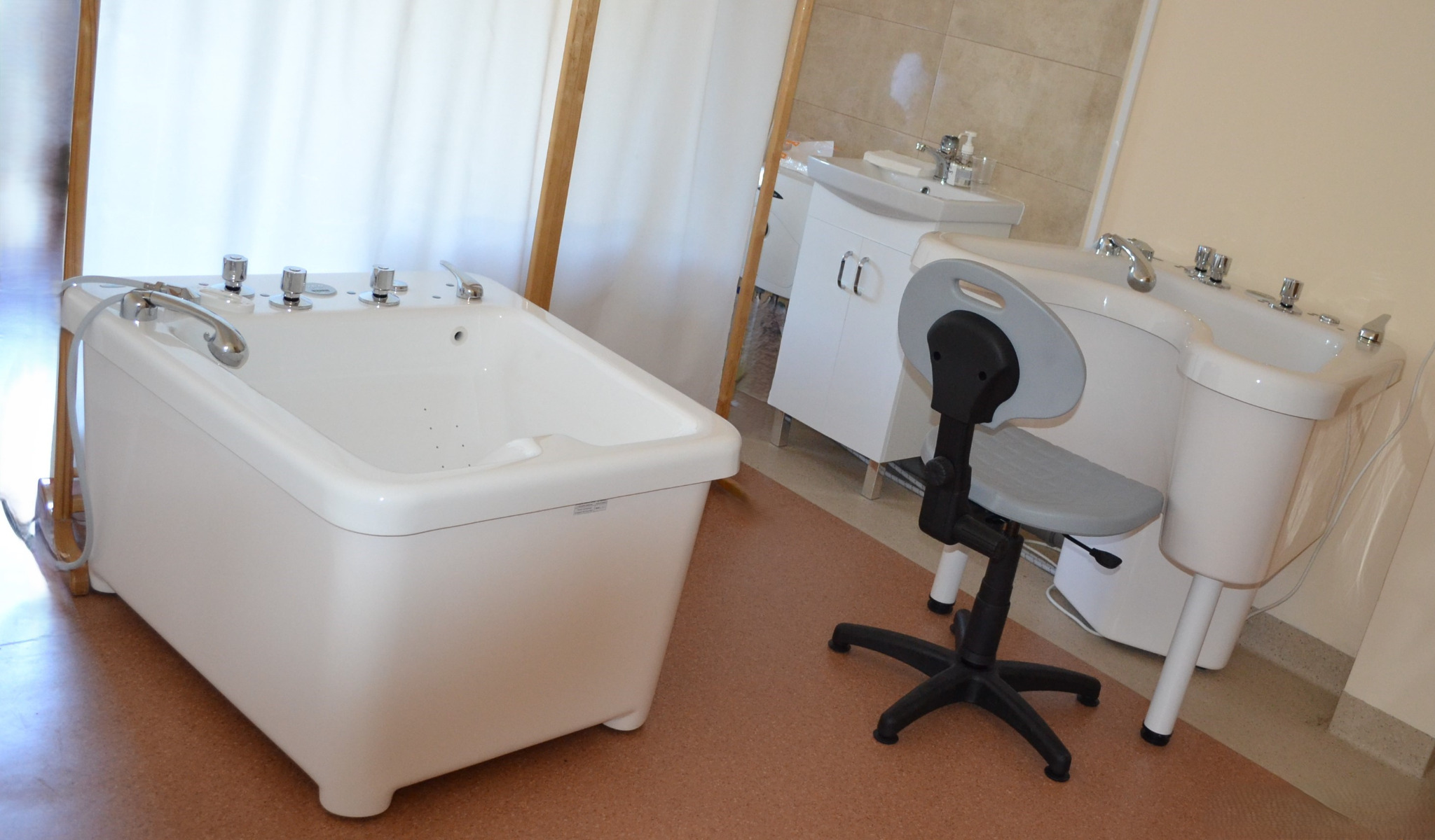 Biała wanna hydromasażowa z chromowanymi pokrętłami i słuchawką prysznicową, obok krzesło z szarą tapicerką, umywalki na postumentach w tle, jasne wnętrze gabinetu.