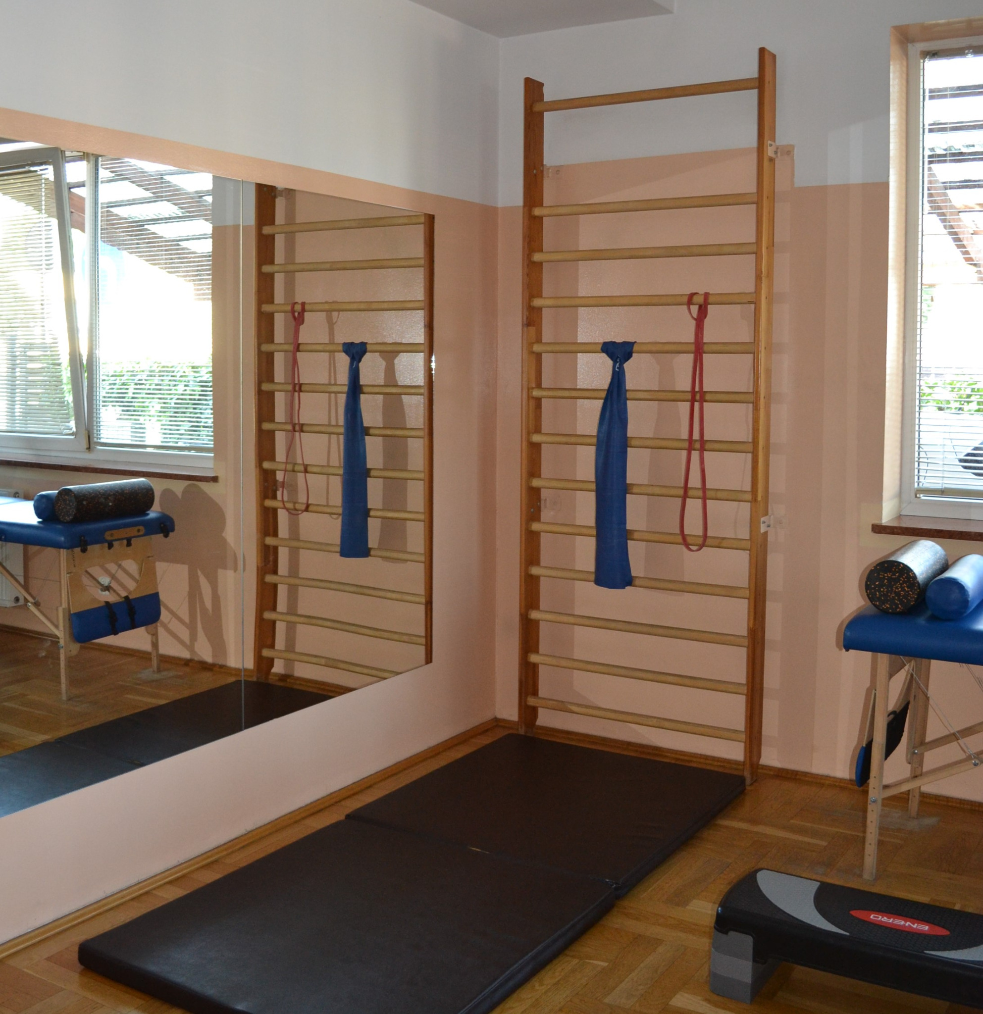 Sala do ćwiczeń z drabinkami gimnastycznymi, matą, stołem do masażu z wałkami i stepem; odbicie w lustrze; naturalne światło z okna.