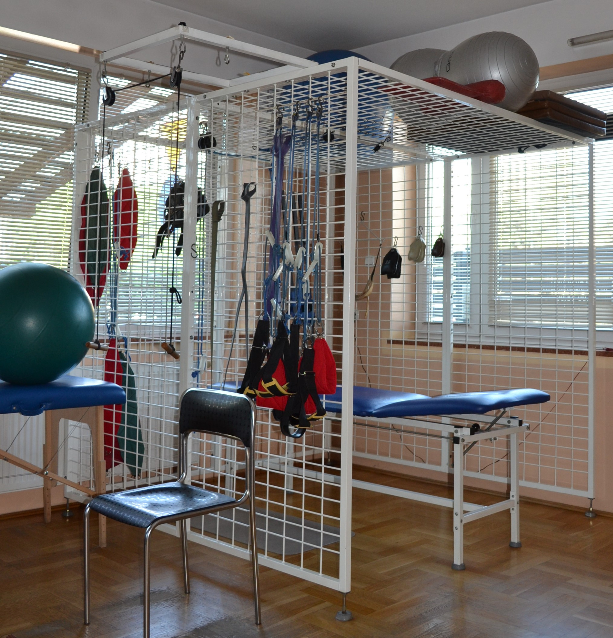 Sala rehabilitacyjna z drabinką rehabilitacyjną, stołem do ćwiczeń i piłkami gimnastycznymi, oświetlona przez okno z żaluzjami.