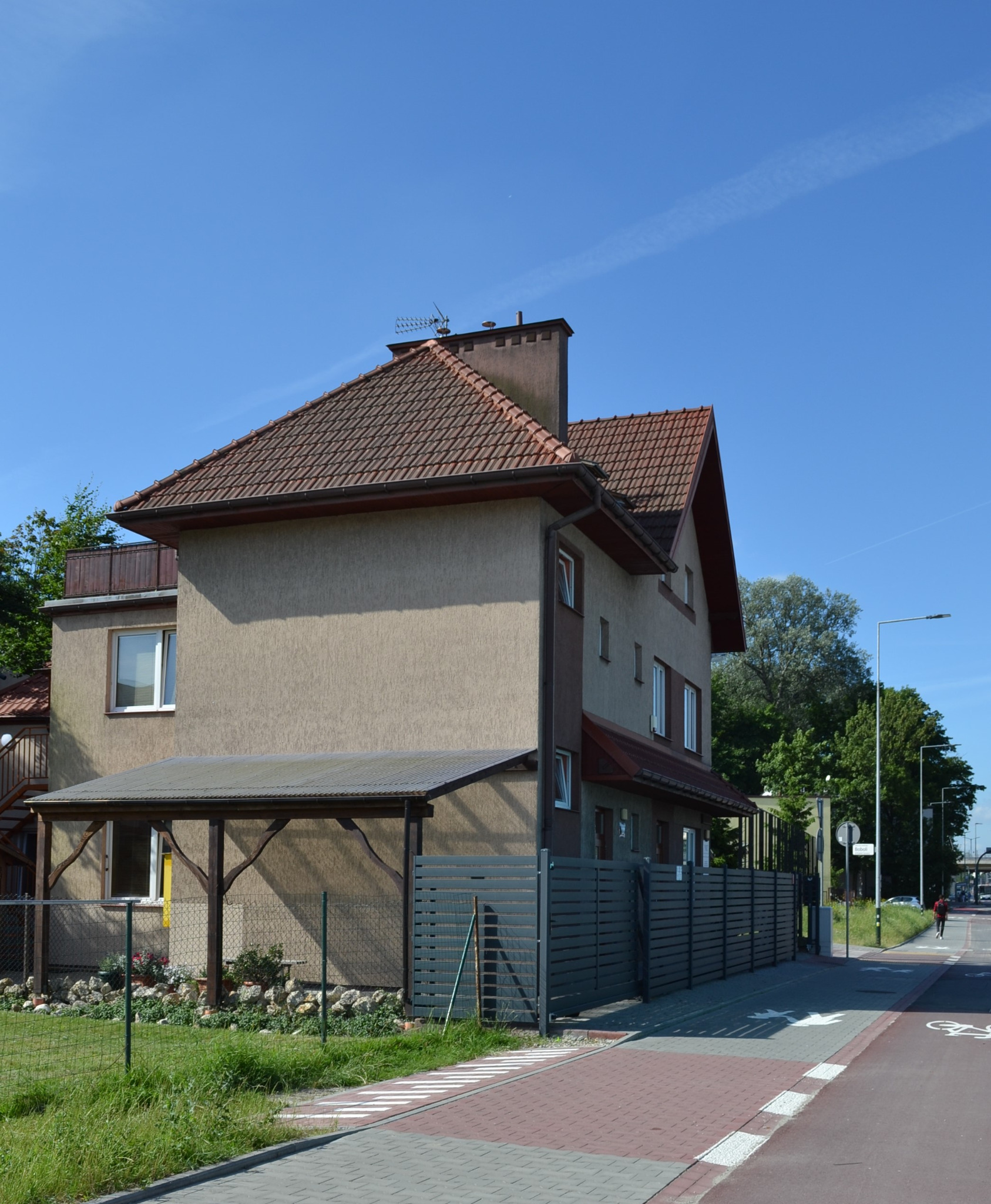 Dwupiętrowy dom z beżową elewacją i brązowym dachem, widoczny z perspektywy ulicy, z przylegającym zadaszeniem i ogrodzeniem z poziomych desek.