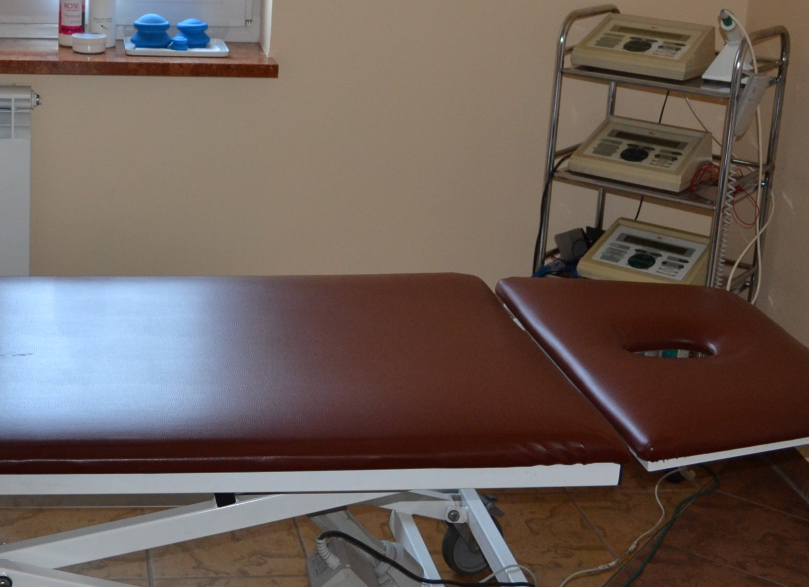 Brązowy stół do terapii z otworem na twarz, obok metalowy wózek z trzema urządzeniami do elektroterapii, w tle okno z niebieskimi bańkami do masażu.