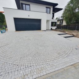 Walintsin Sych - Podjazd do nowoczesnego domu wyłożony szarą kostką granitową w okrągły wzór, z widocznymi schodami z kostki prowadzącymi w stronę ogrodu.