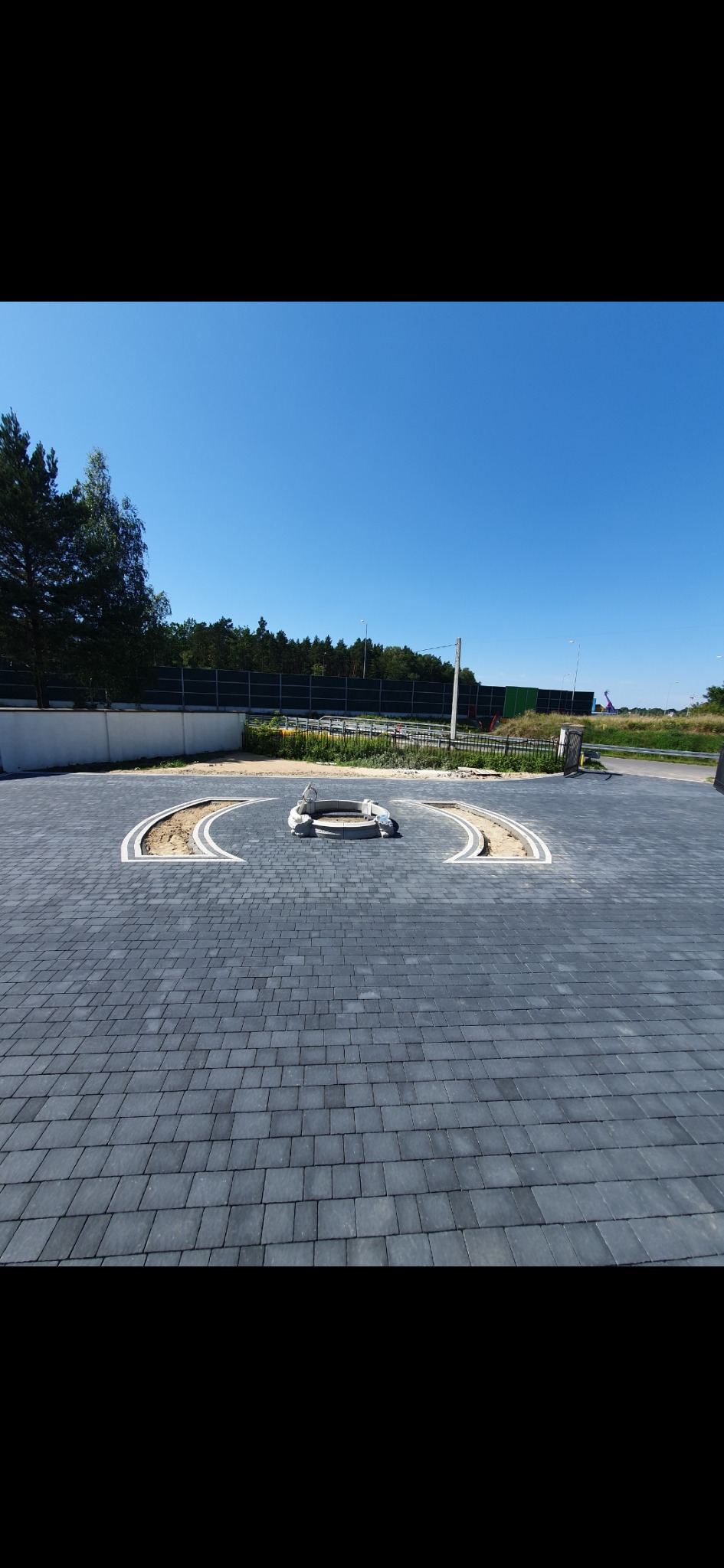 Nawierzchnia z ciemnoszarej kostki brukowej ułożonej na dużej powierzchni, z centralnie umieszczonym elementem dekoracyjnym w postaci okręgu z piasku i białego obrzeża. W tle widoczny pas zieleni...