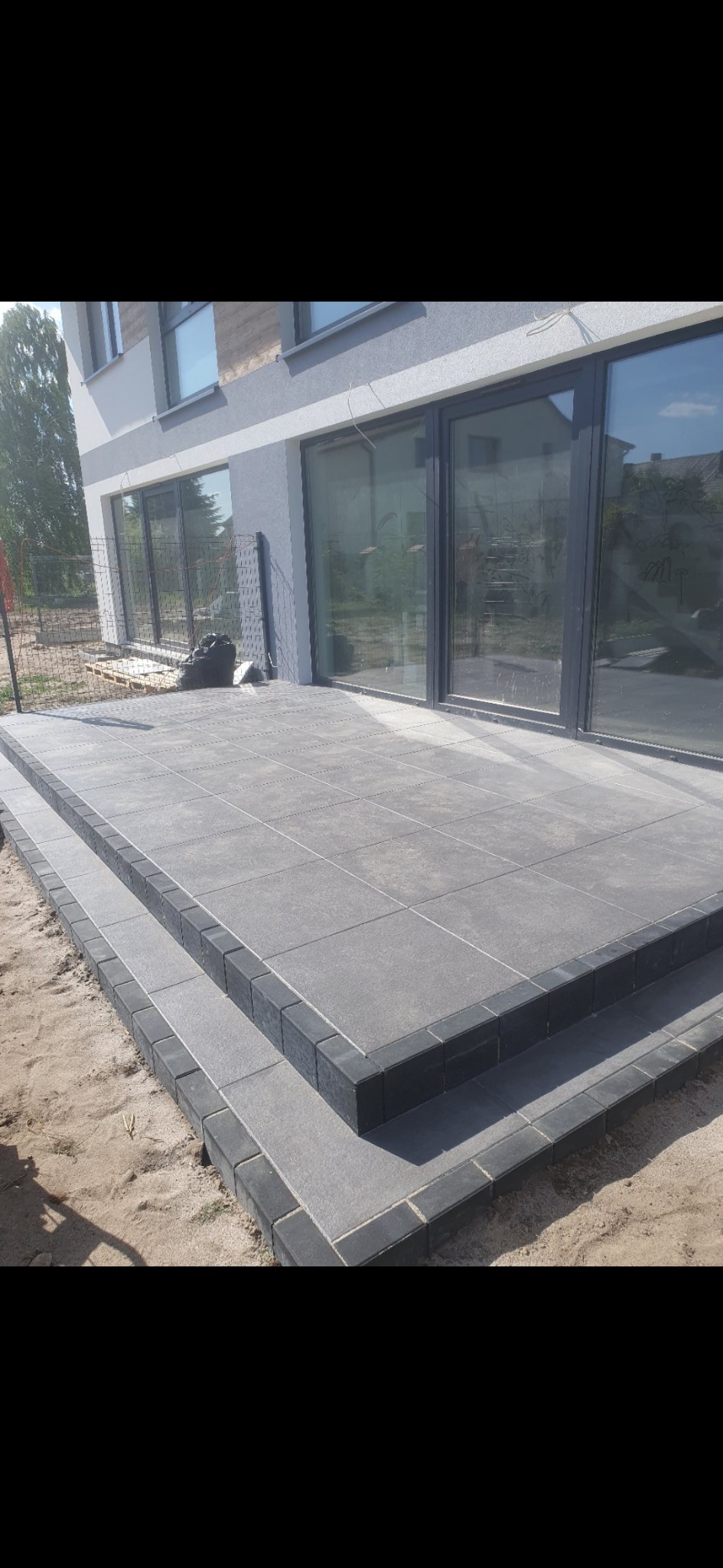Nowoczesny taras z szarych płyt betonowych z ciemnoszarą obwódką z kostki, prowadzący do dużych, przesuwnych drzwi tarasowych w nowym domu, widoczne ślady piasku po pracach brukarskich.