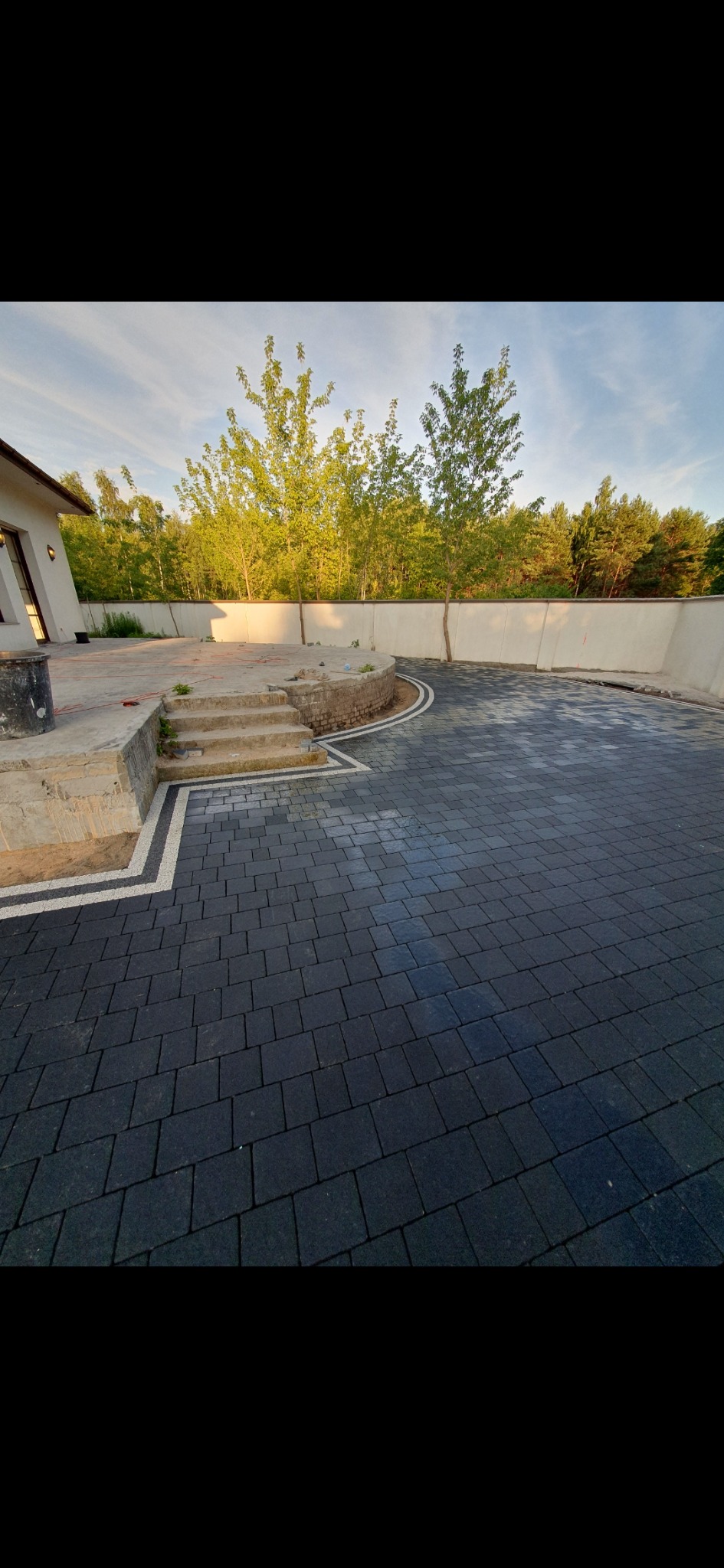 Ułożona, grafitowa kostka brukowa z jasną obwódką, sąsiadująca z betonowymi schodami i przygotowanym terenem pod dalsze prace wykończeniowe, na tle białego muru i drzew.