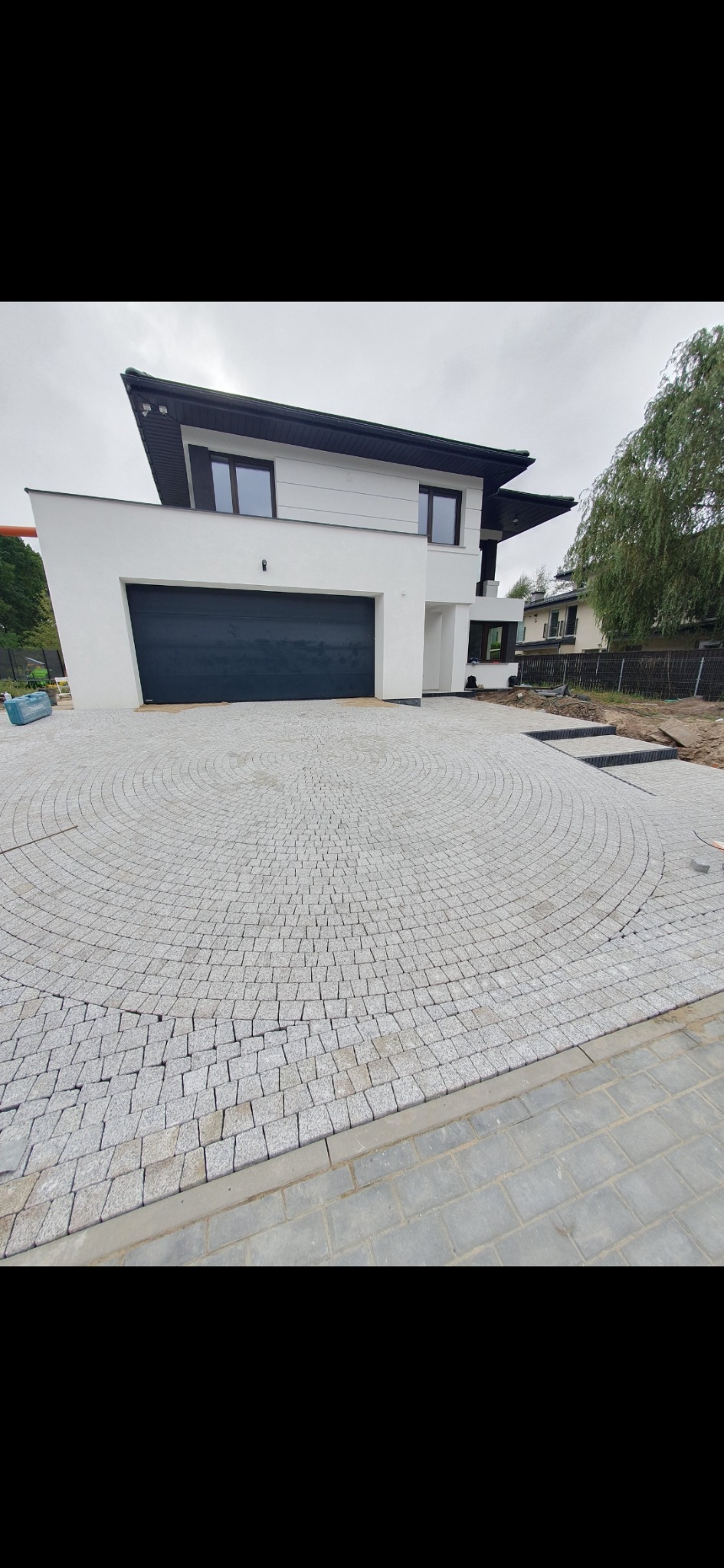 Podjazd do nowoczesnego domu wyłożony szarą kostką granitową w okrągły wzór, z widocznymi schodami z kostki prowadzącymi w stronę ogrodu.