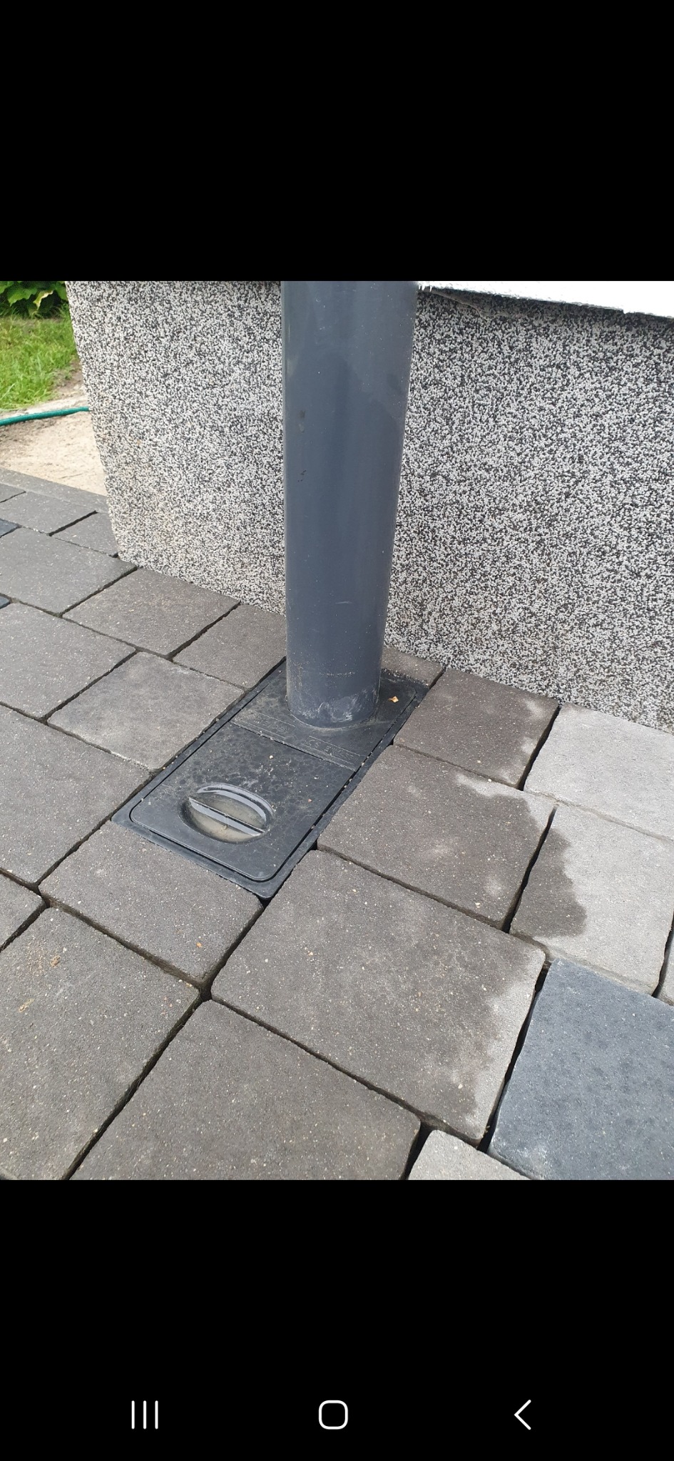 Szara kostka brukowa ułożona wokół szarej rury spustowej z czarną studzienką kanalizacyjną przy ścianie budynku o szarej, ziarnistej elewacji.