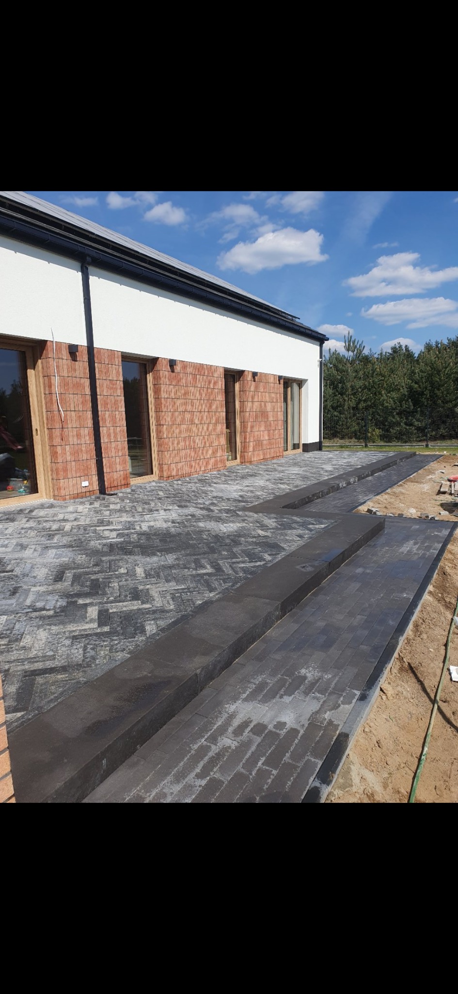 Ułożona, szara kostka brukowa na podjeździe domu z ceglaną elewacją, widoczne przygotowane krawężniki i piasek.