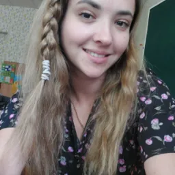 Portret uśmiechniętej kobiety z blond włosami, zaplecionymi w warkocz z ozdobnymi srebrnymi gumkami, ubrana w czarną sukienkę w różowe kwiaty, na tle zielonej tablicy i ściany w geometryczne wzory.