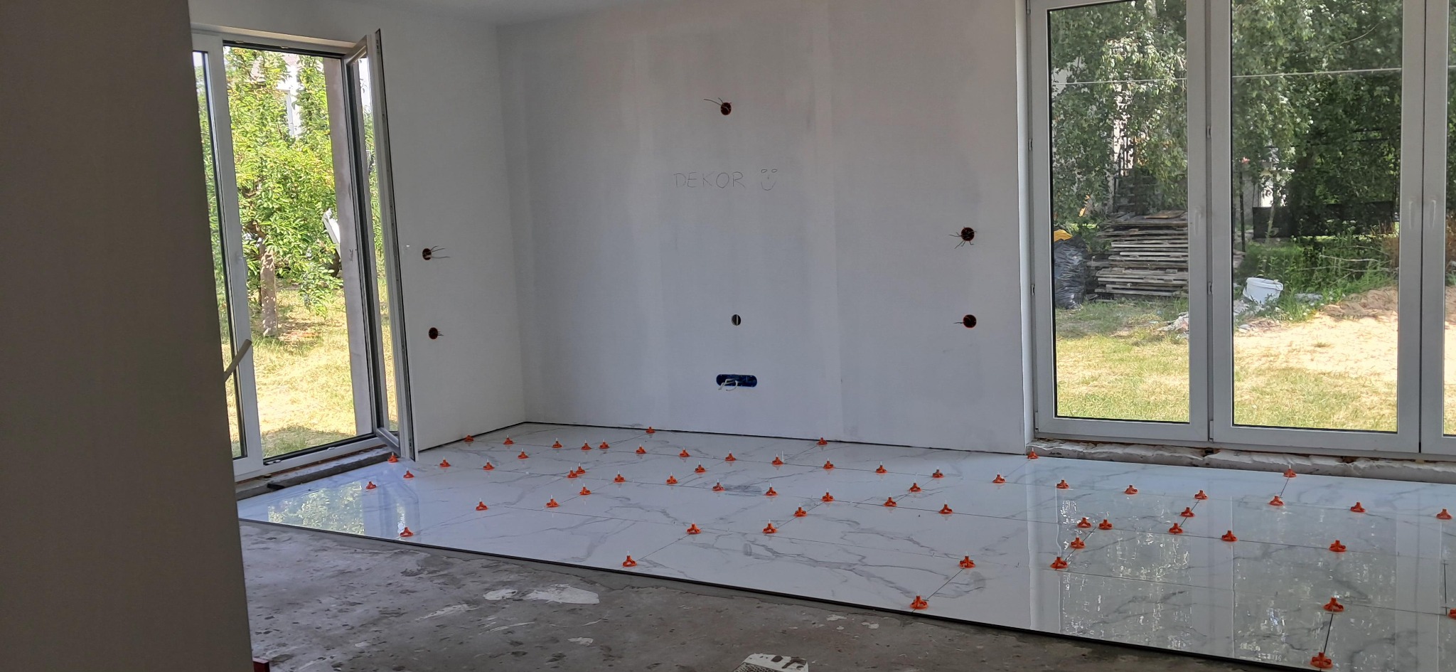 Układanie białych płytek podłogowych z marmurowym wzorem, z użyciem pomarańczowych klipsów poziomujących. Widoczne otwory na gniazdka w ścianie.